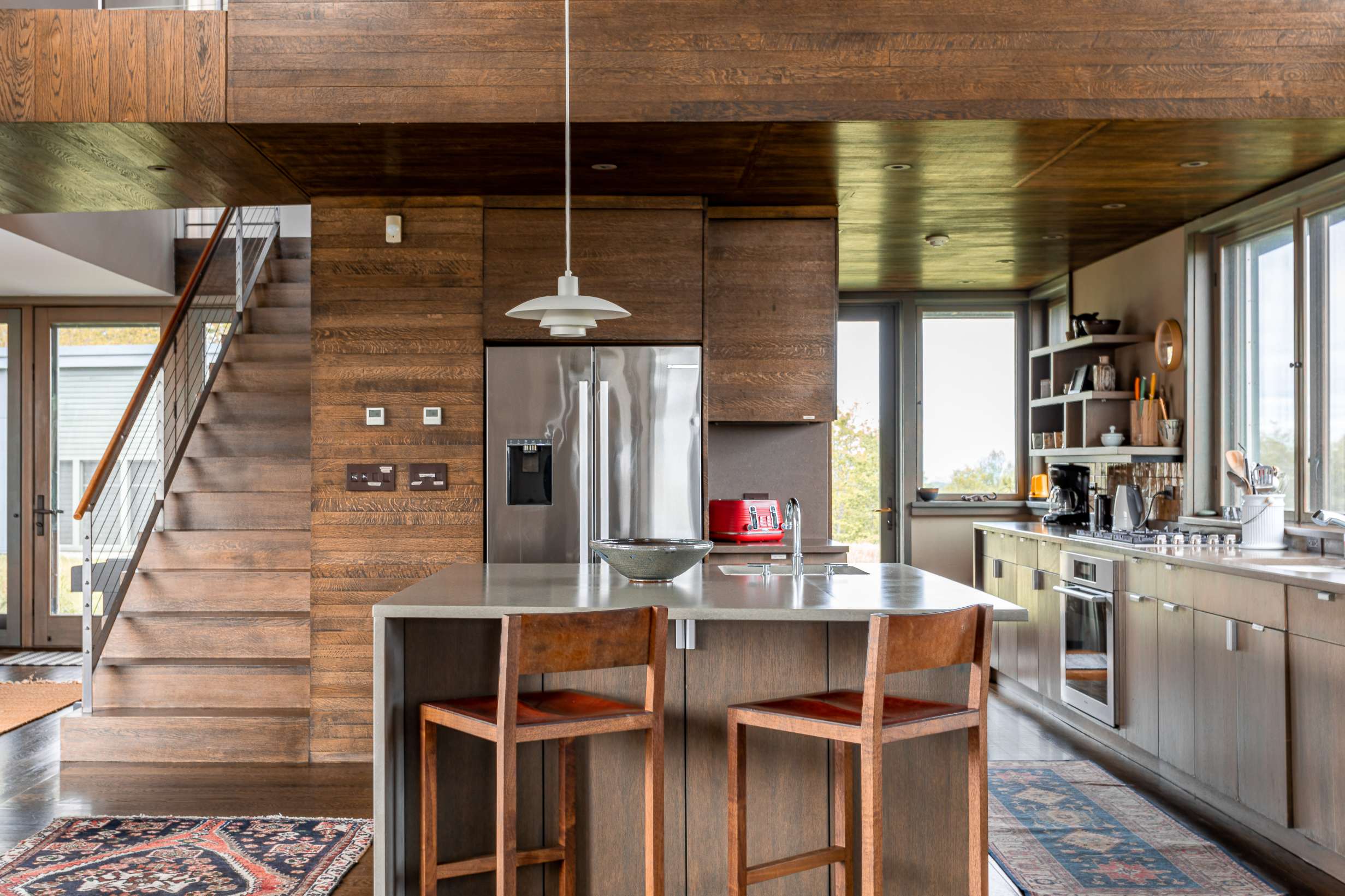 Modern kitchen with wood cabinetry, stainless steel appliances, and a large island with bar stools, featuring a striking wood-paneled wall and staircase leading upstairs.
