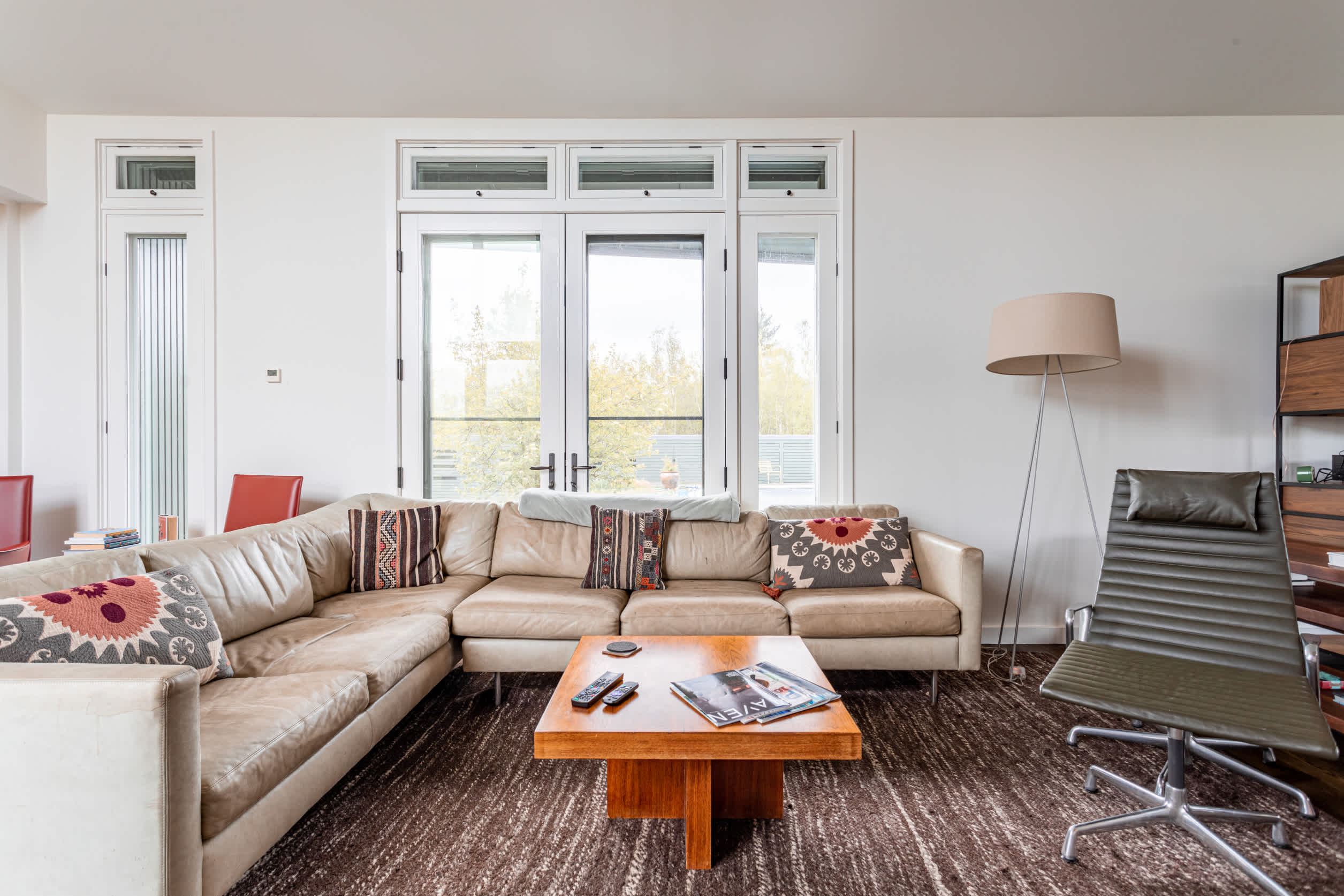 A bright living room features a large L-shaped leather sofa, a wooden coffee table, and a modern floor lamp, with multiple windows and glass doors providing natural light and views of the outdoor landscape.
