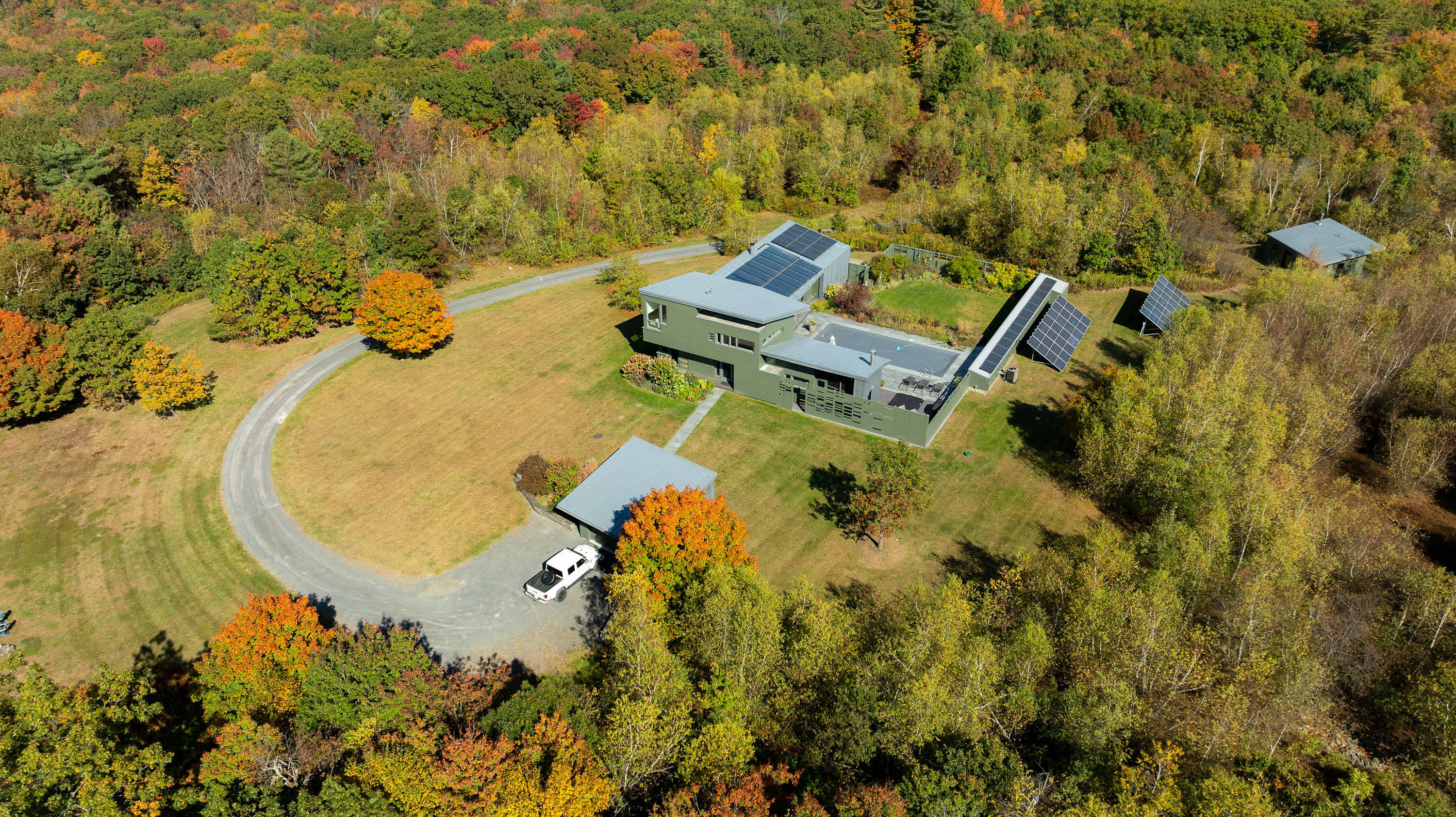 An aerial view of the property during autumn, showing the green house with solar panels on the roof, surrounded by a mix of green and colorful fall foliage. A circular driveway and additional solar panels are visible on the grounds.