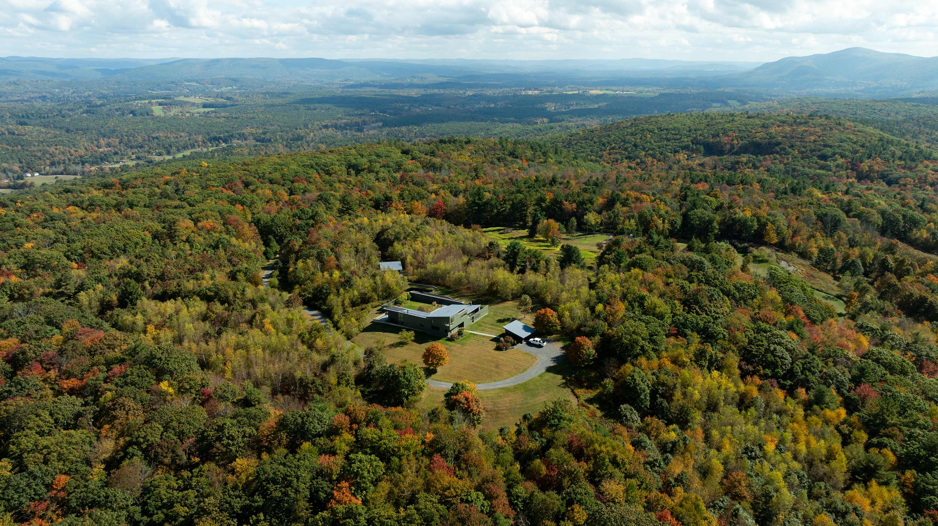 A sprawling modern house with a dark facade is situated on a hilltop, surrounded by a vast expanse of autumn-colored trees and distant mountains under a partly cloudy sky, showcasing its secluded location.