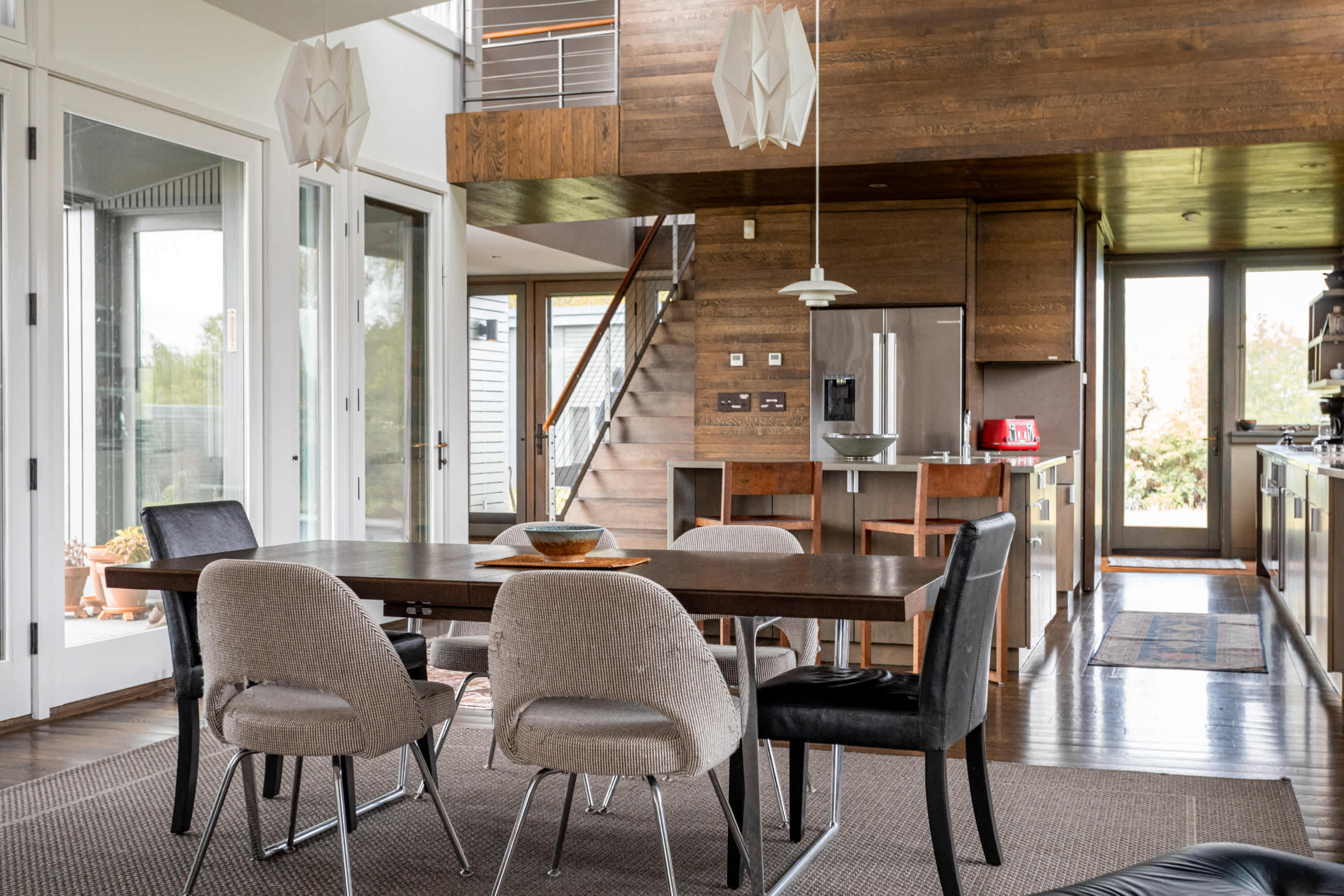 Open-concept dining area with a dark wood table and mixed chairs on a light grey rug, offering a view into the modern kitchen and staircase beyond.