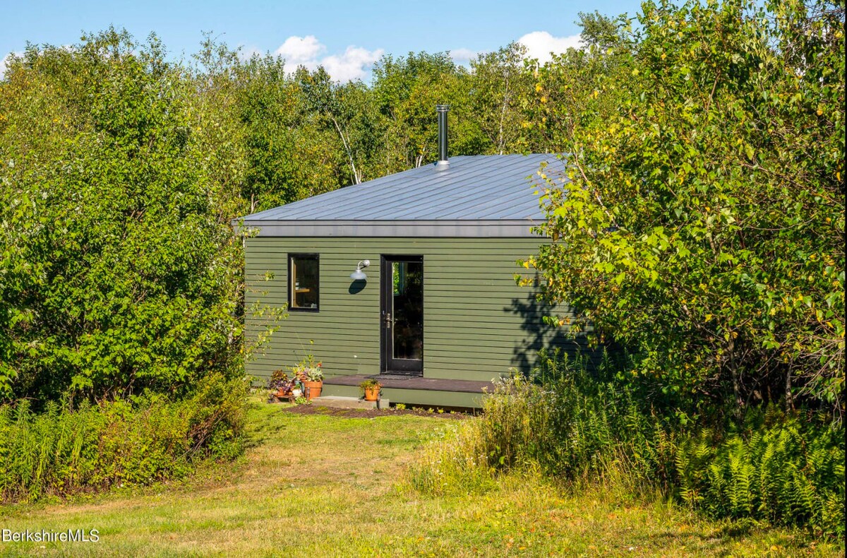 A small, modern green-sided building with a gray metal roof, nestled among lush green trees and foliage. A single door and window are visible on the front, with a small porch area.