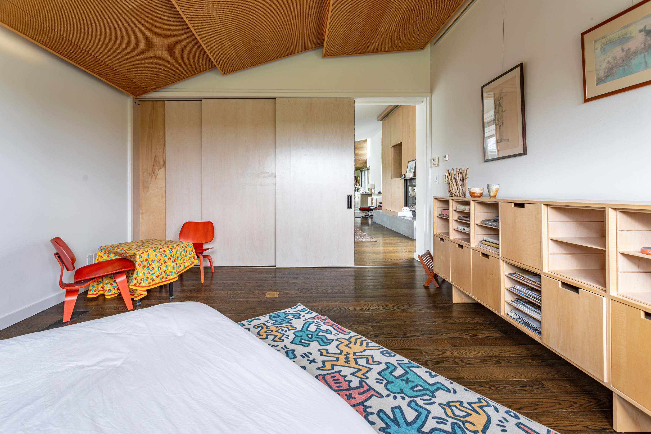 View of a contemporary bedroom with dark wood floors and a colorful rug, looking towards a large sliding door entrance. A light wood storage cabinet is visible, along with a small red table and chairs.