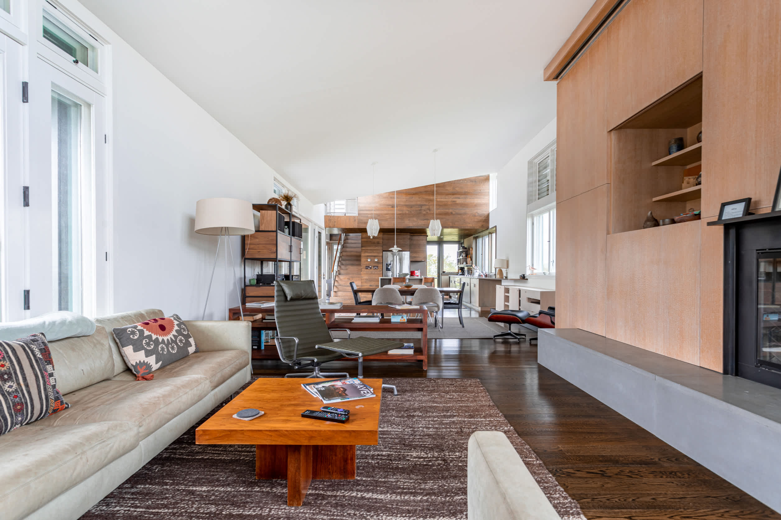Open-concept living room with a light-colored sofa, wooden coffee table, and a modern fireplace with a light wood surround, overlooking the dining and kitchen areas.
