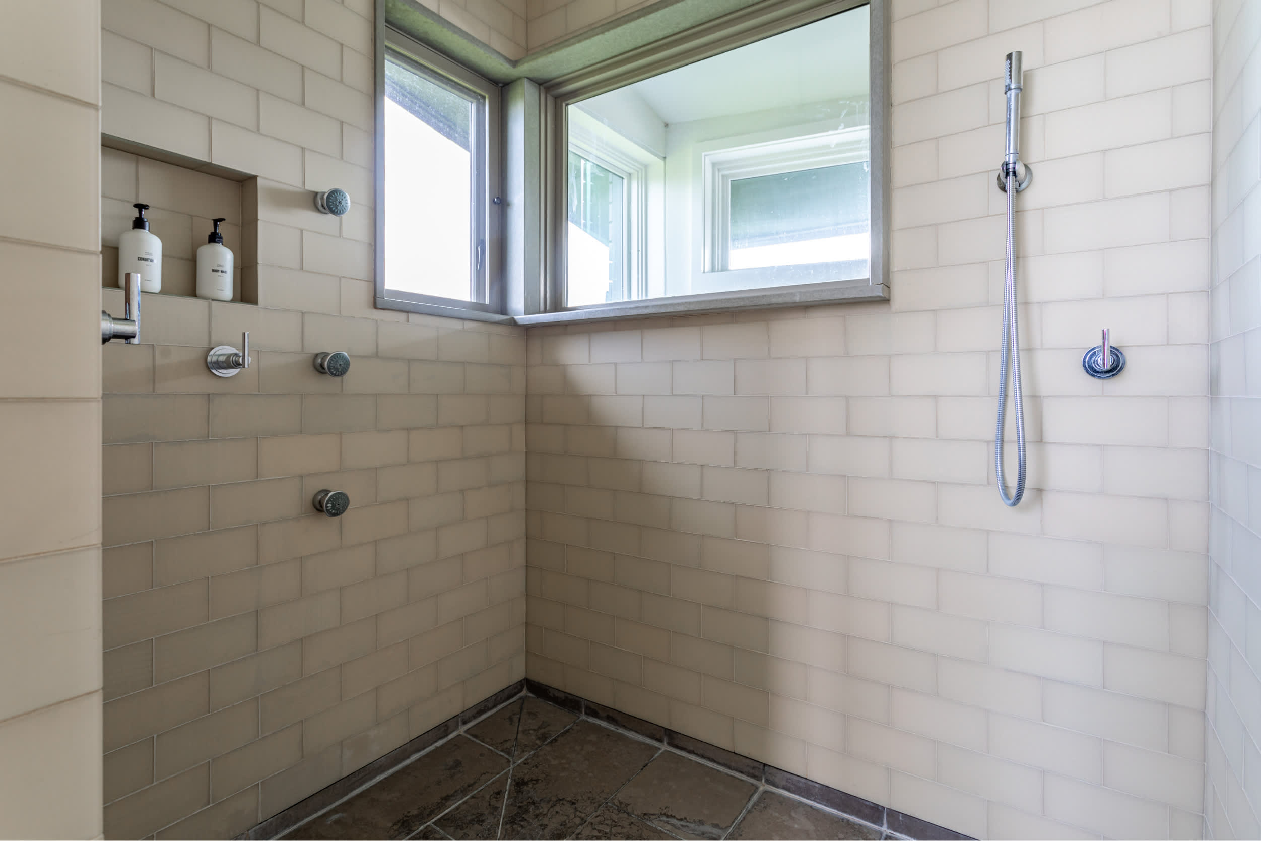 Spacious walk-in shower with light beige subway tiles, multiple shower heads, and a built-in niche, featuring a window for natural light.