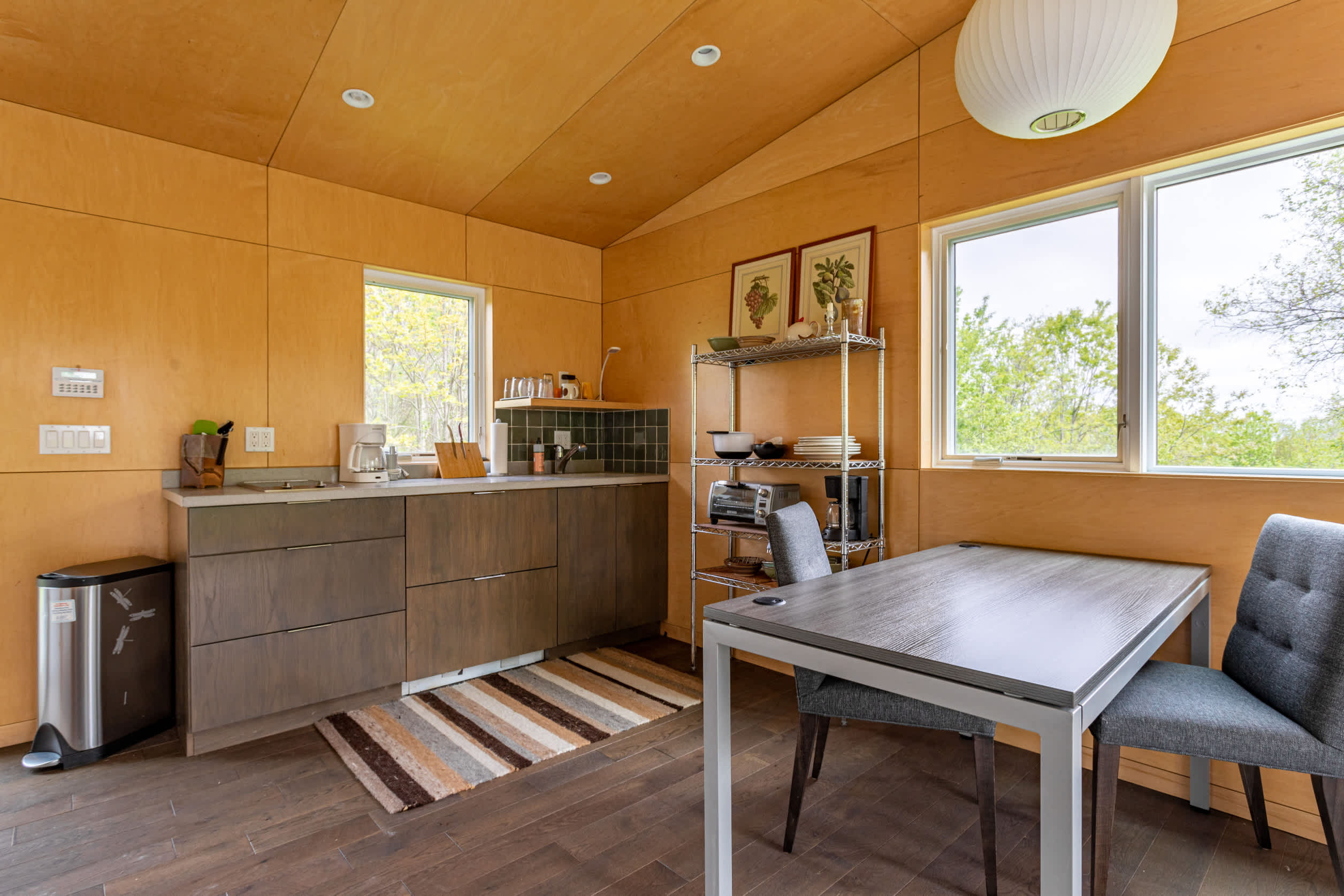 Detailed view of a compact kitchen area with dark flat-panel cabinets, a stainless steel shelf, and a light wood countertop. The walls and ceiling are covered in light wood paneling, and a dining table is nearby.