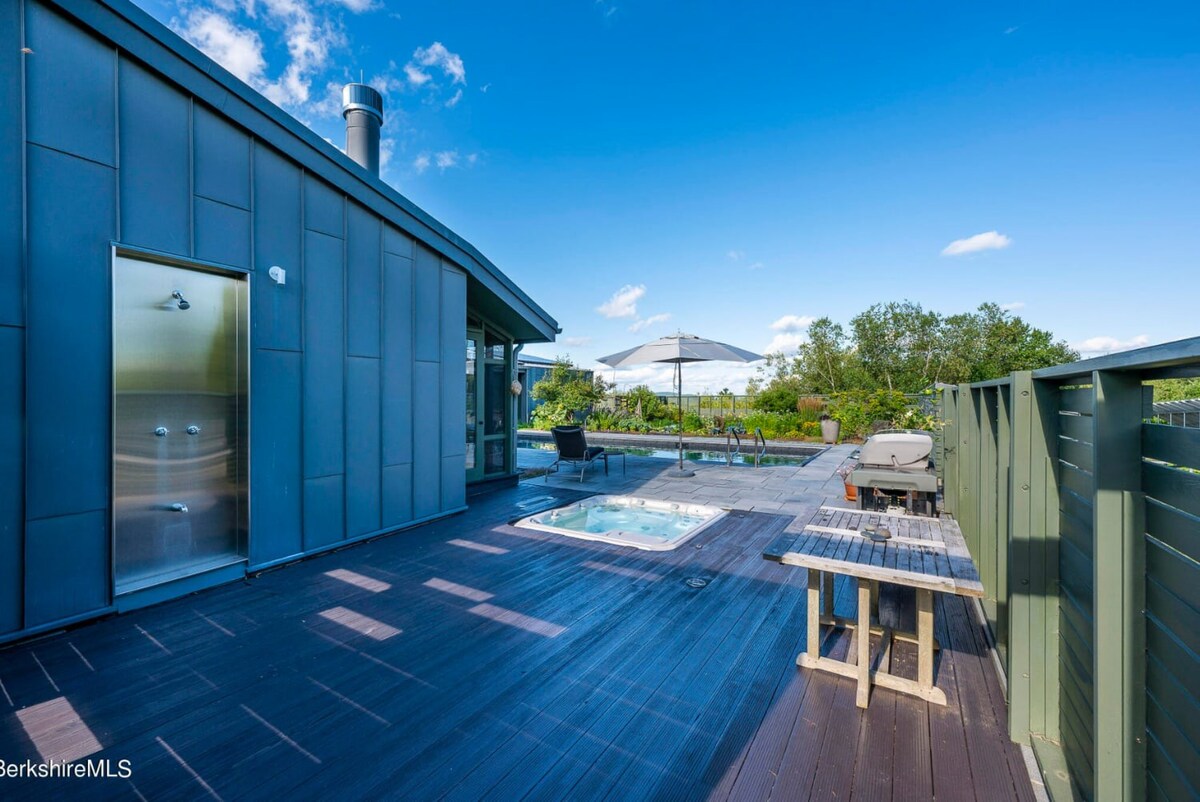 A spacious outdoor deck featuring a built-in hot tub, a BBQ grill, and an outdoor table. The deck is made of dark wood, and the area is surrounded by a fence and lush greenery under a clear blue sky.