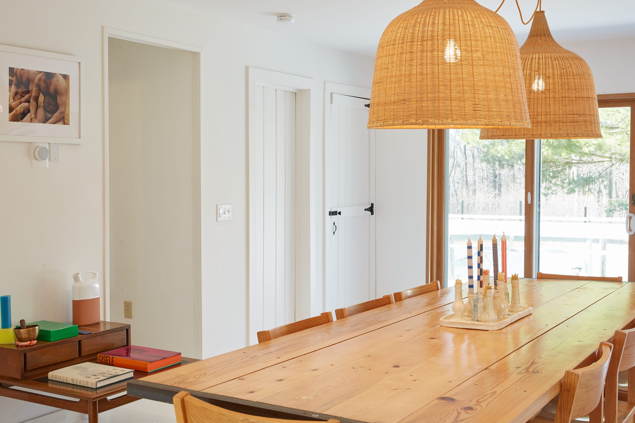 Close-up of the dining table with decorative candles, showing the light wooden chairs and the two large woven pendant lights. White doors and sliding glass doors leading to the deck are visible in the background.