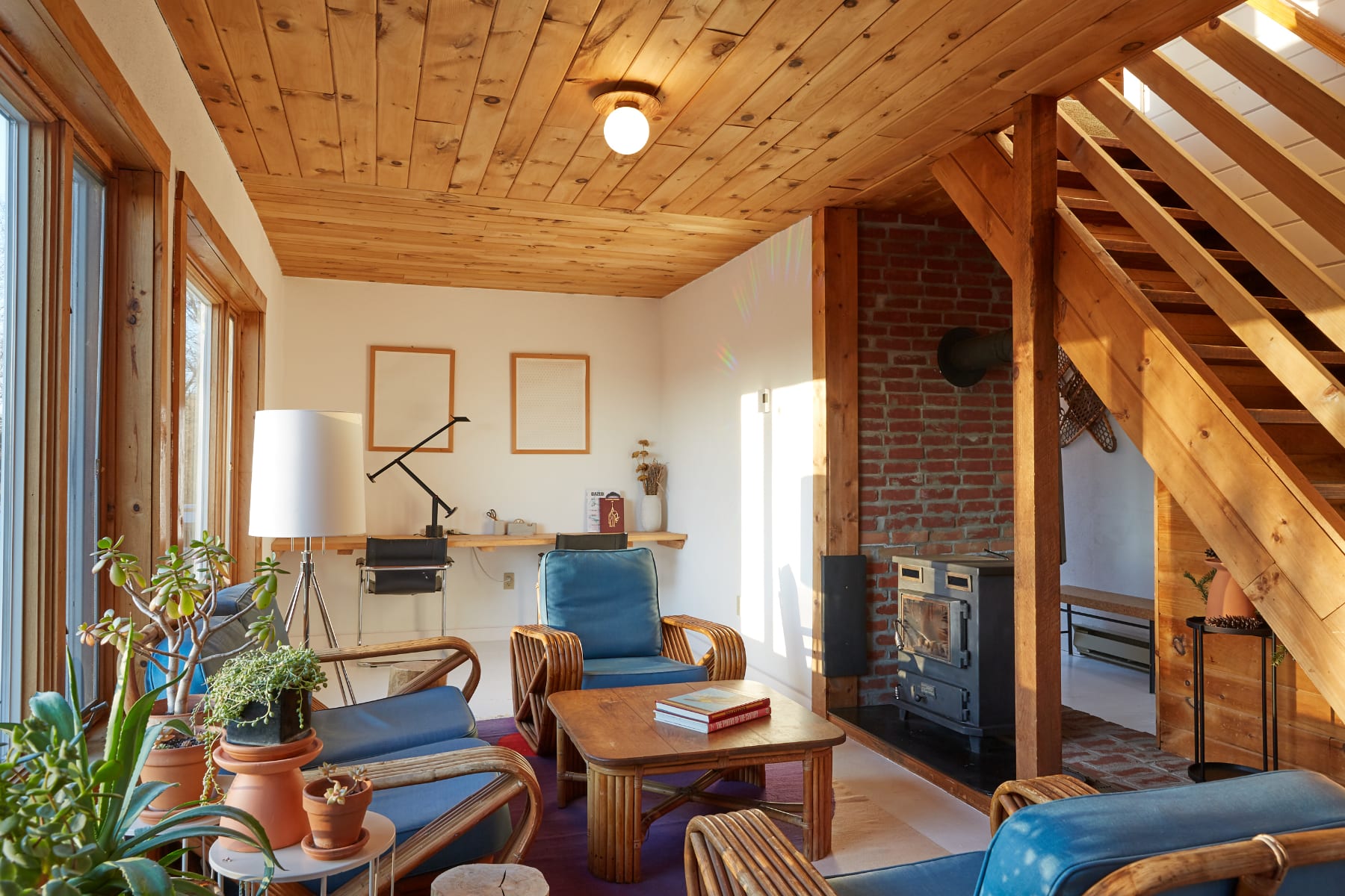 Cozy living room with a wooden plank ceiling, large windows, and a brick fireplace with a wood-burning stove. Rattan furniture with blue cushions and various potted plants create a warm, inviting atmosphere.