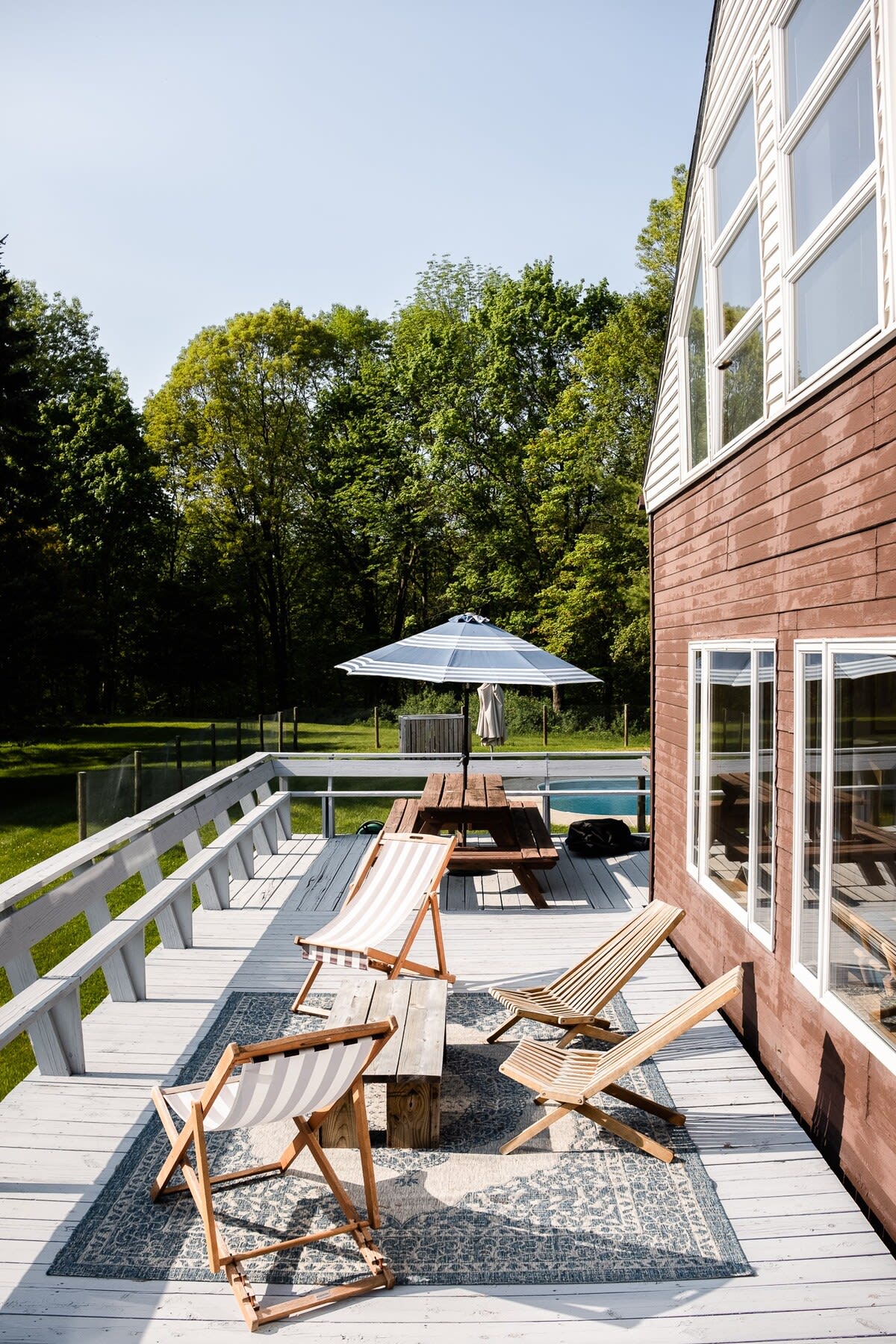 A spacious outdoor deck with light gray wooden planks, featuring a patterned rug, several wooden lounge chairs, and a picnic table with a blue and white striped umbrella. The brown-sided house with large windows is visible on the right.