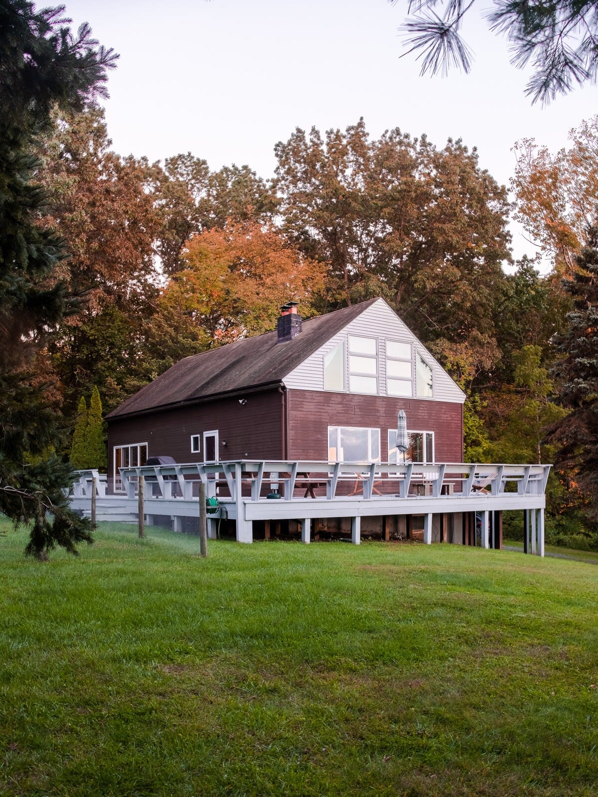 A dark brown, two-story cabin with a light gray deck wraps around the front, set on a grassy hill surrounded by autumn trees. The upper level features large, multi-pane windows.