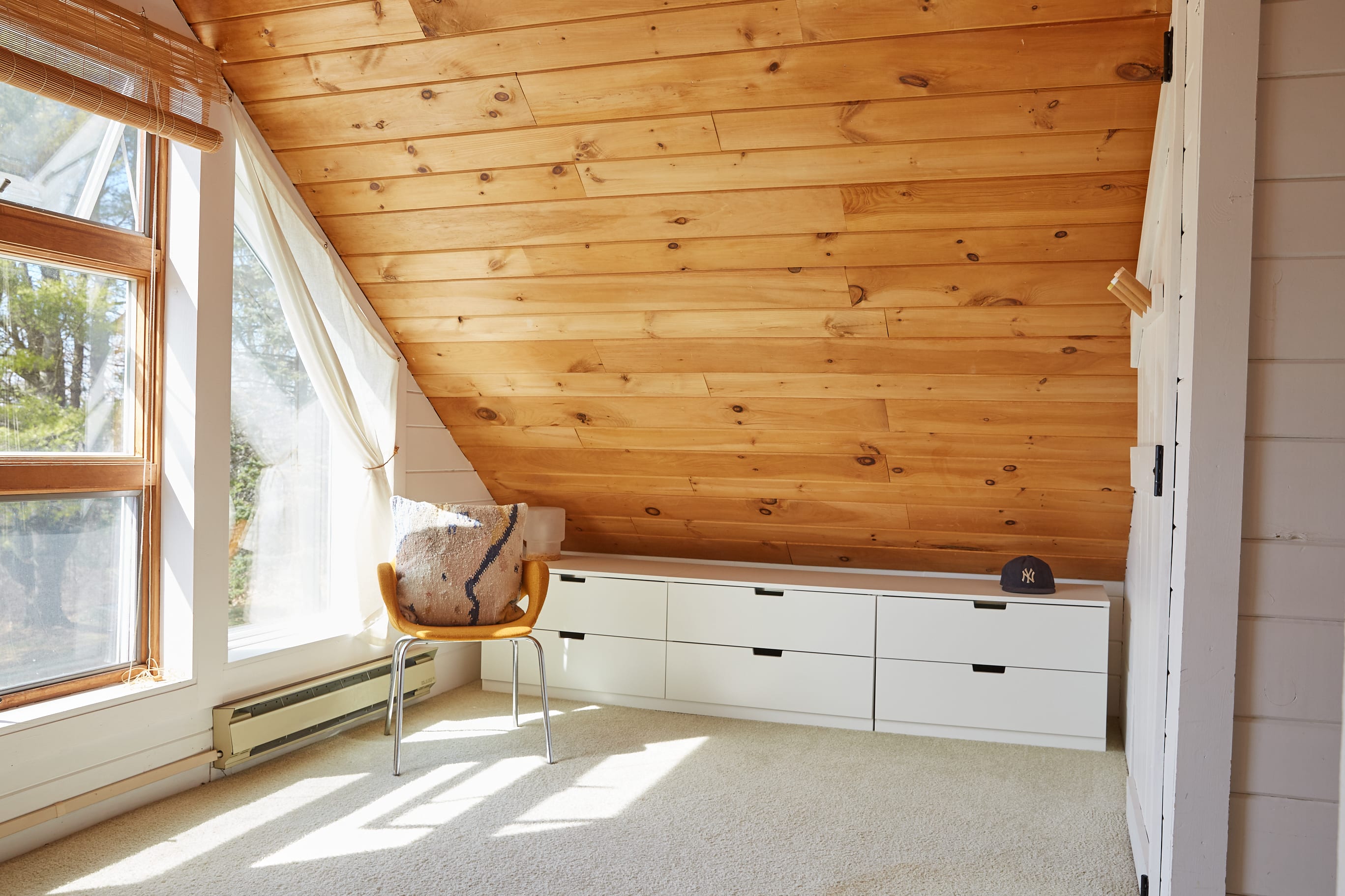 Attic space with a sloped wooden plank ceiling and large windows, featuring a long white dresser or storage unit along the wall. A single chair and a baseball cap are on the dresser, with sunlight streaming onto the carpeted floor.