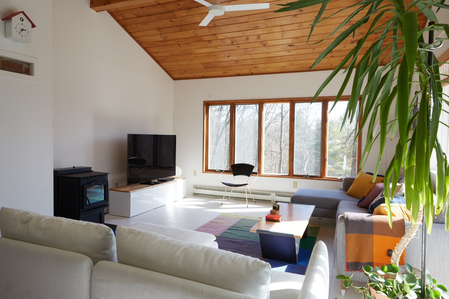 Bright living room with white walls, a vaulted wooden ceiling, and light-colored flooring. A large gray sectional sofa, a black wood-burning stove, and a TV are visible, with large windows overlooking trees.