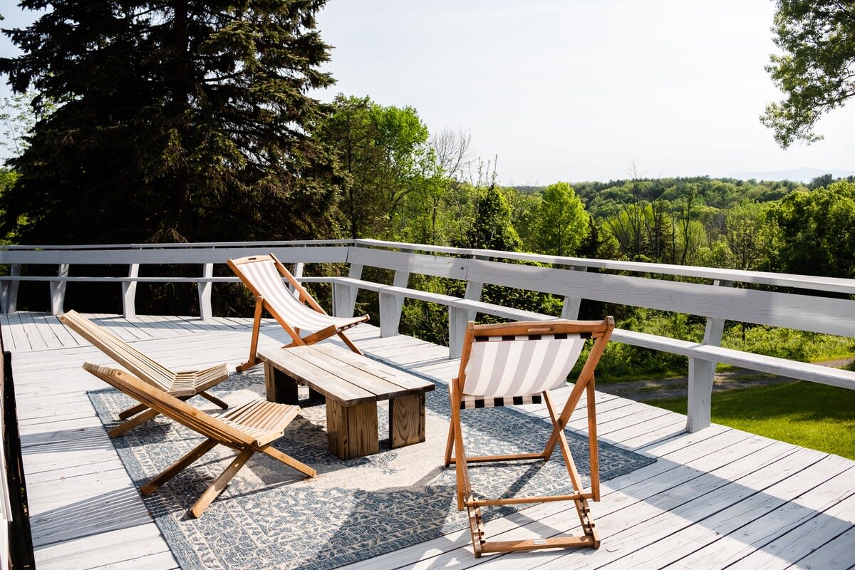An expansive white wooden deck featuring a patterned outdoor rug and three stylish wooden lounge chairs, providing panoramic views of the verdant landscape and distant hills.