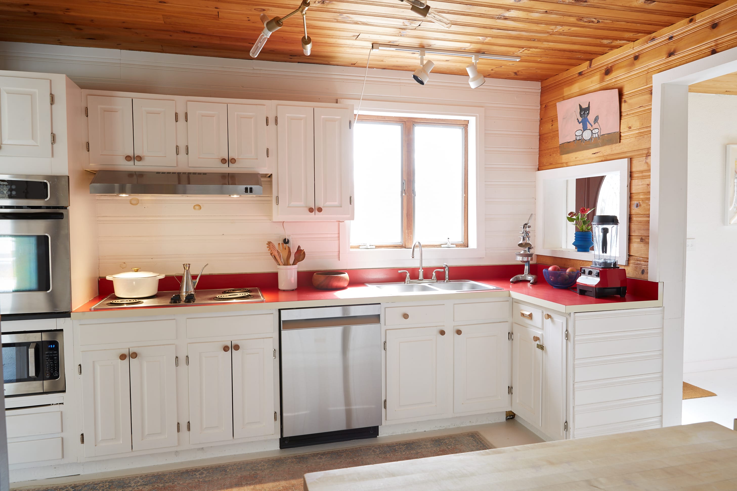 Well-lit kitchen with white shaker-style cabinets, vibrant red countertops, and a double sink under a window. The room features a warm wooden ceiling and shiplap accent wall, with stainless steel appliances.