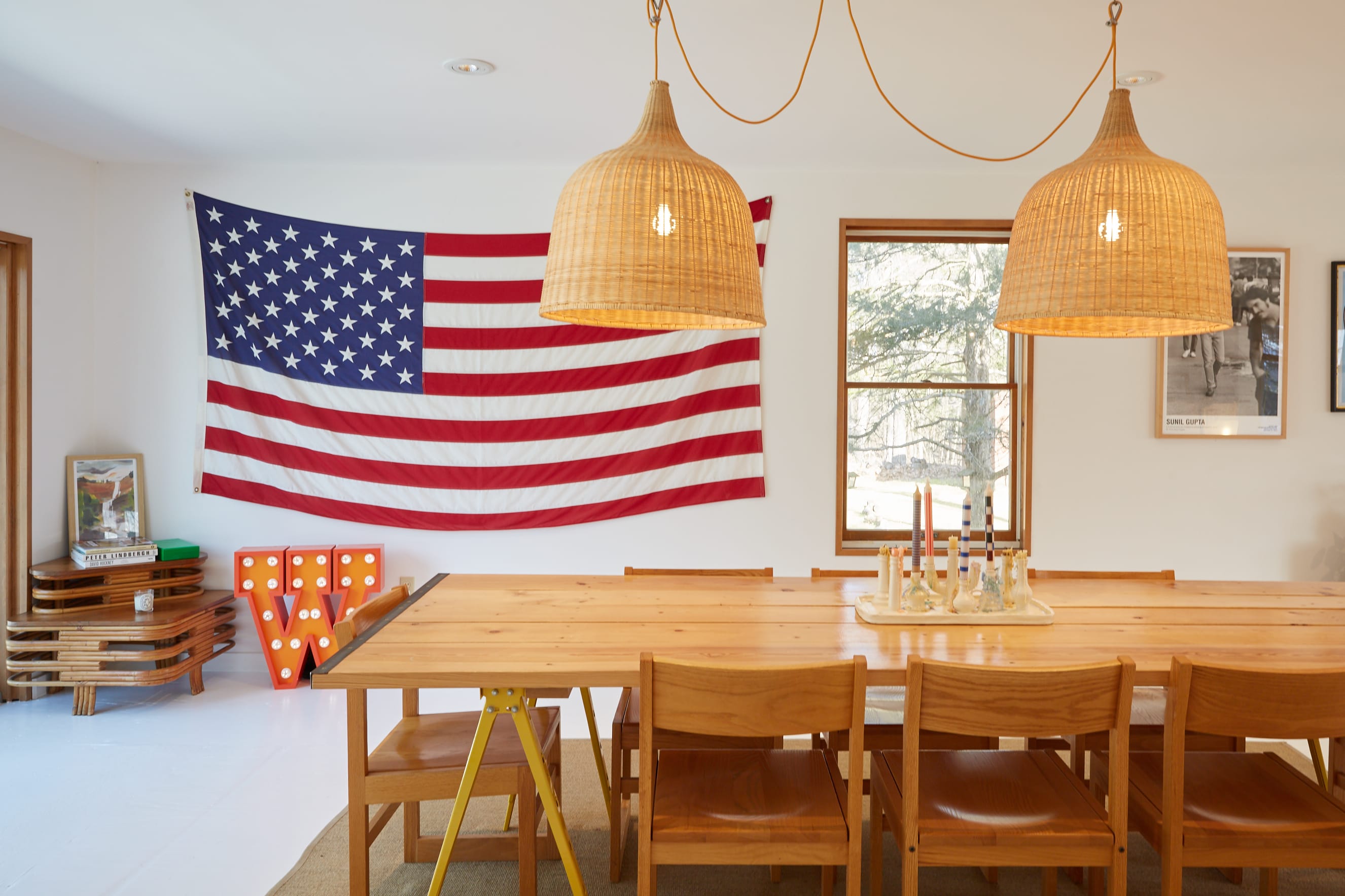 A spacious dining room featuring a long wooden table with matching chairs, illuminated by two woven pendant lights, and accented by a large American flag displayed prominently on the wall.