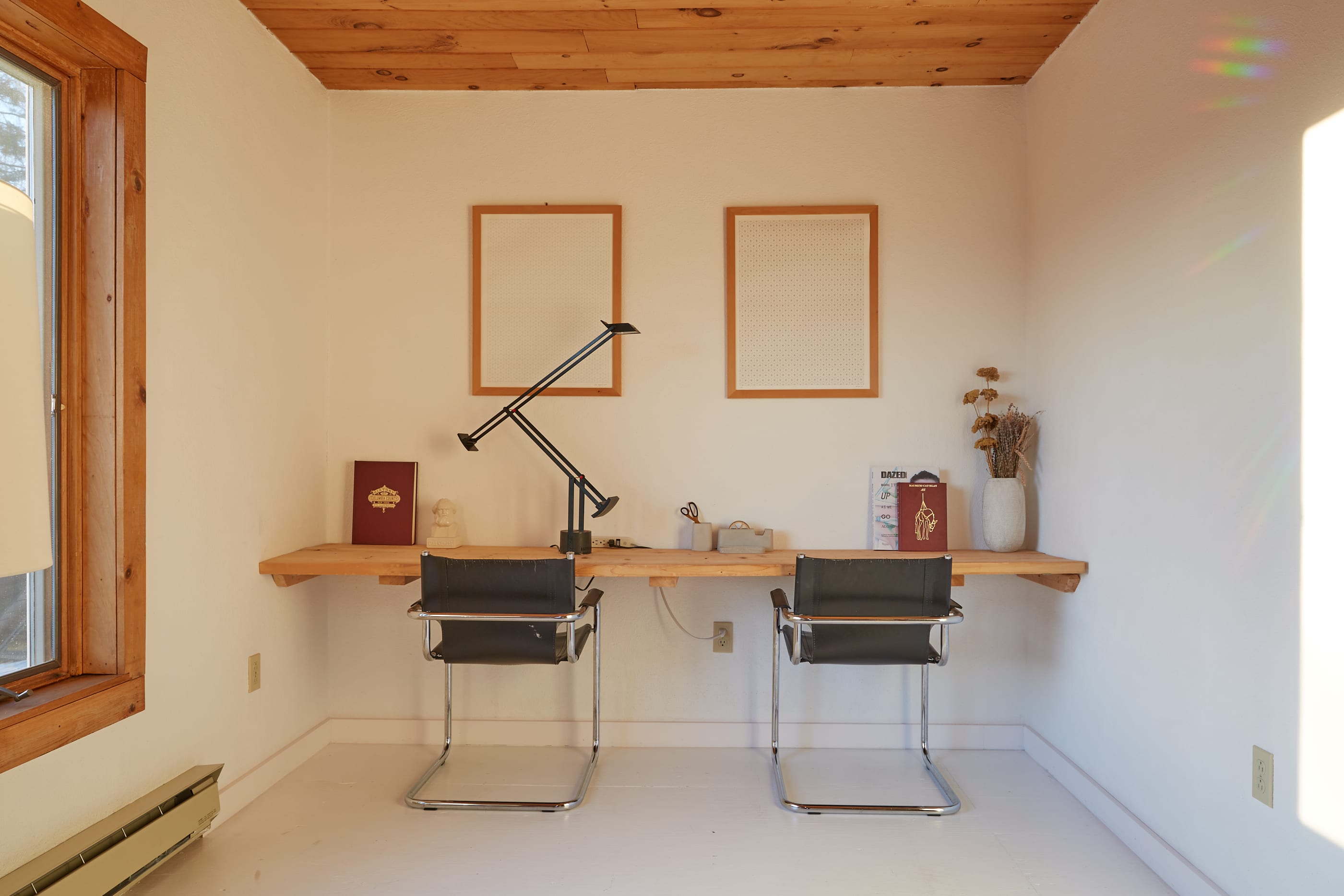 Dedicated workspace area within the living room, featuring a long wooden floating desk with two black chairs and a modern desk lamp. Two framed artworks adorn the white wall, with natural light streaming in from a nearby window.