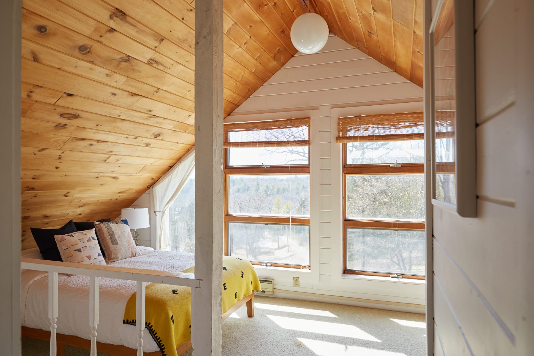 Bright loft bedroom featuring a sloped wooden plank ceiling, white shiplap walls, and large windows offering a view of the surrounding trees. A bed with a yellow blanket and patterned pillows is visible behind a white railing.