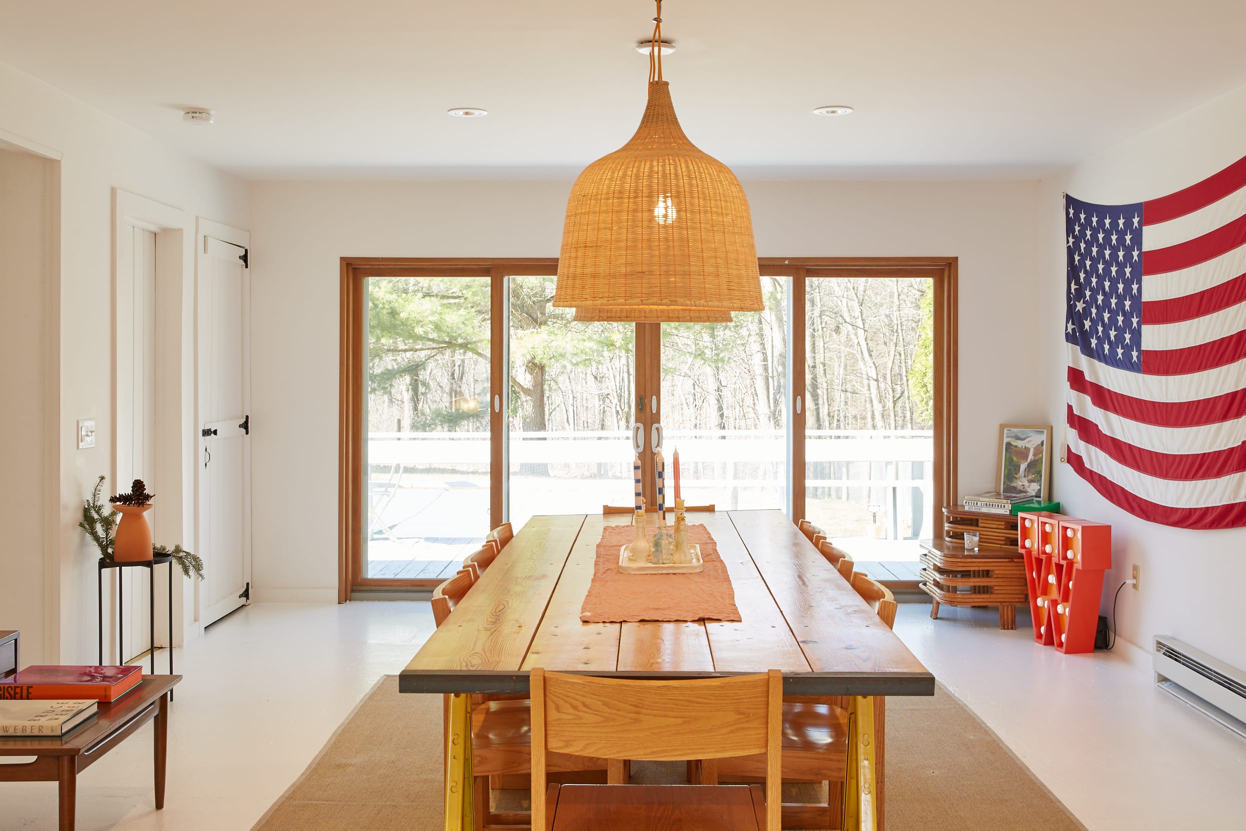 Spacious dining room with a long wooden table and matching chairs, illuminated by a large woven pendant light. Sliding glass doors open to a deck, and a large American flag hangs on the wall.