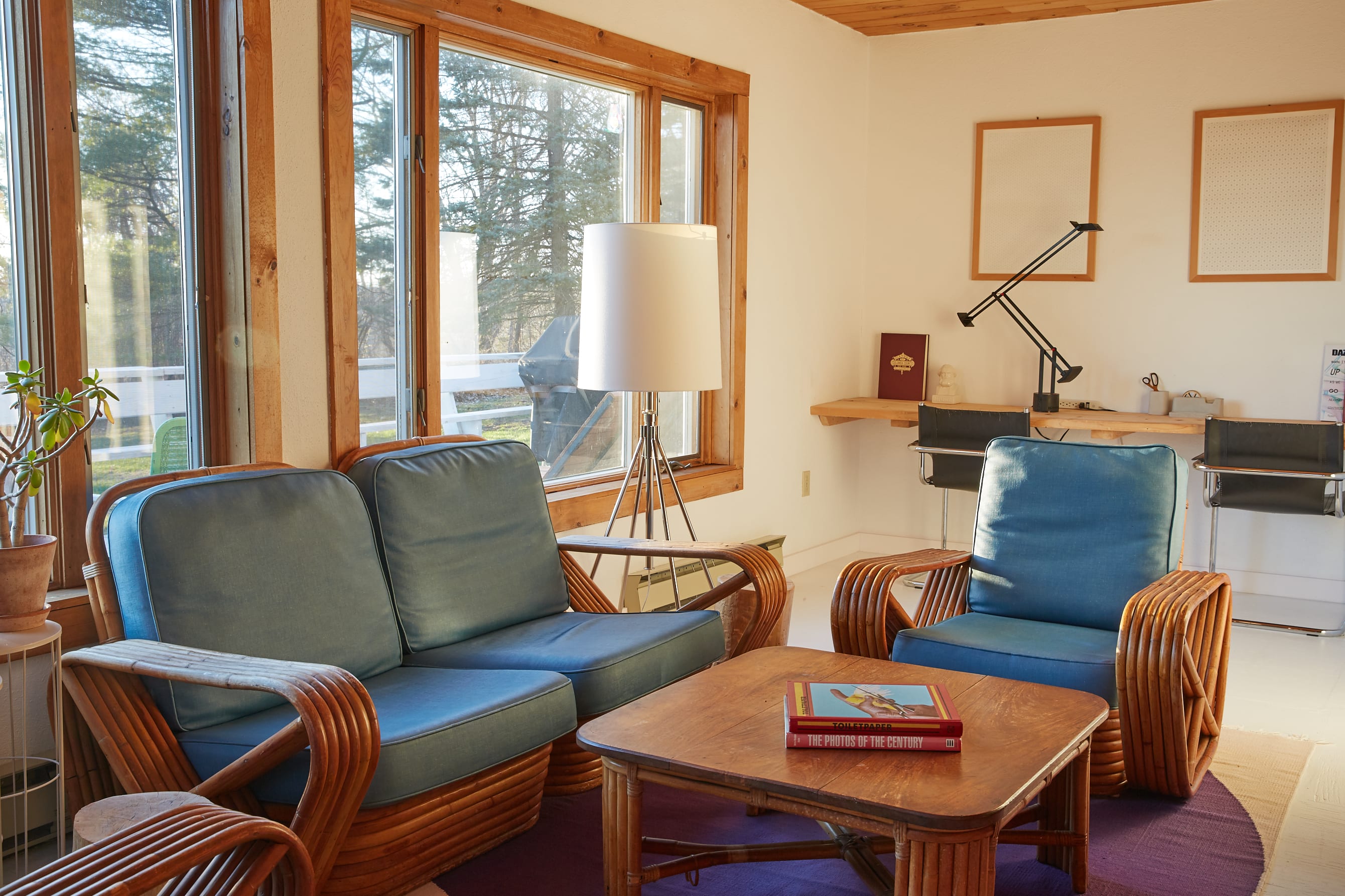 Bright living room with large windows and a wooden plank ceiling, furnished with rattan seating featuring blue cushions and a wooden coffee table. A floor lamp and a dedicated workspace with a floating desk are also visible.