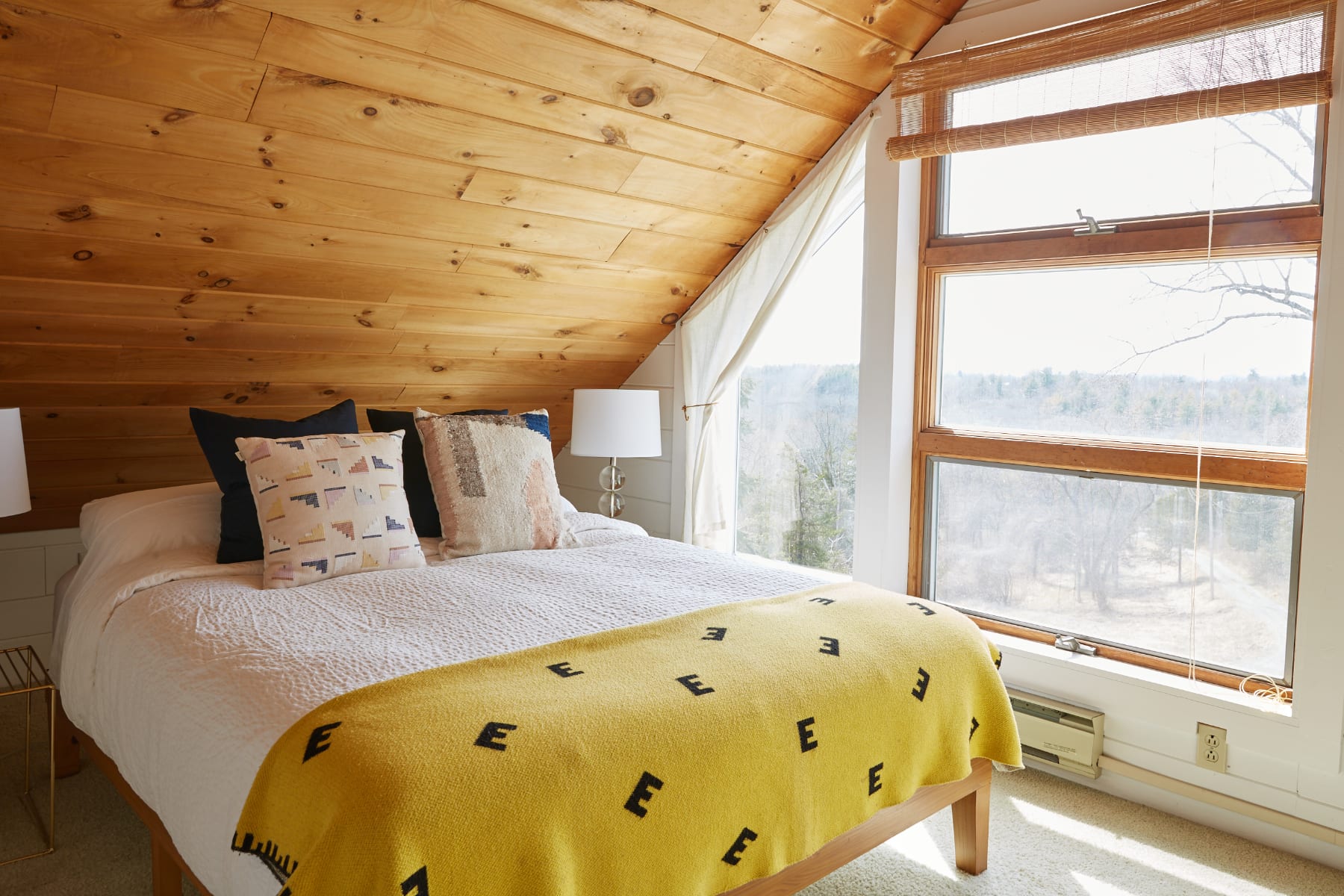 Bright attic bedroom with a sloped wooden plank ceiling and large windows providing expansive views of the surrounding trees. A bed with a yellow blanket and decorative pillows is positioned against the wall, with a nightstand and lamp.