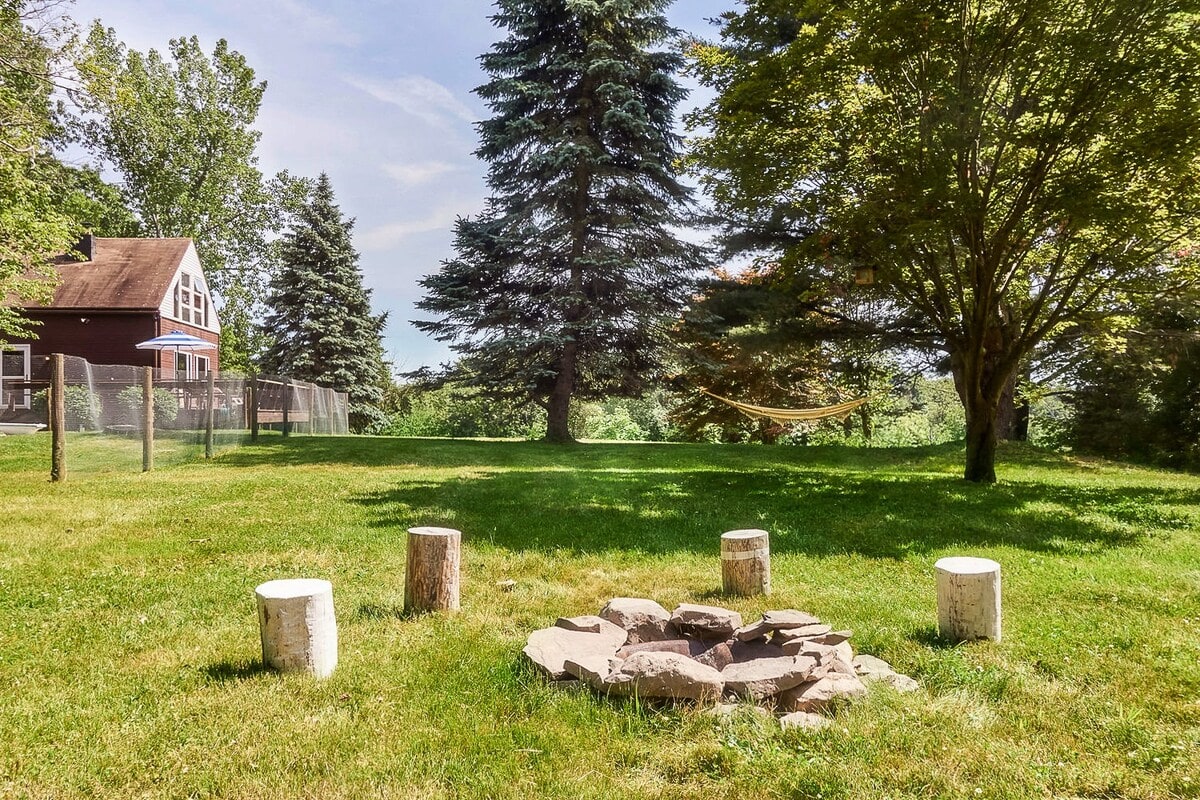 An outdoor fire pit area with a circular stone ring and four rustic log seats, situated on a well-maintained grassy lawn surrounded by mature trees, with a yellow hammock visible in the background.