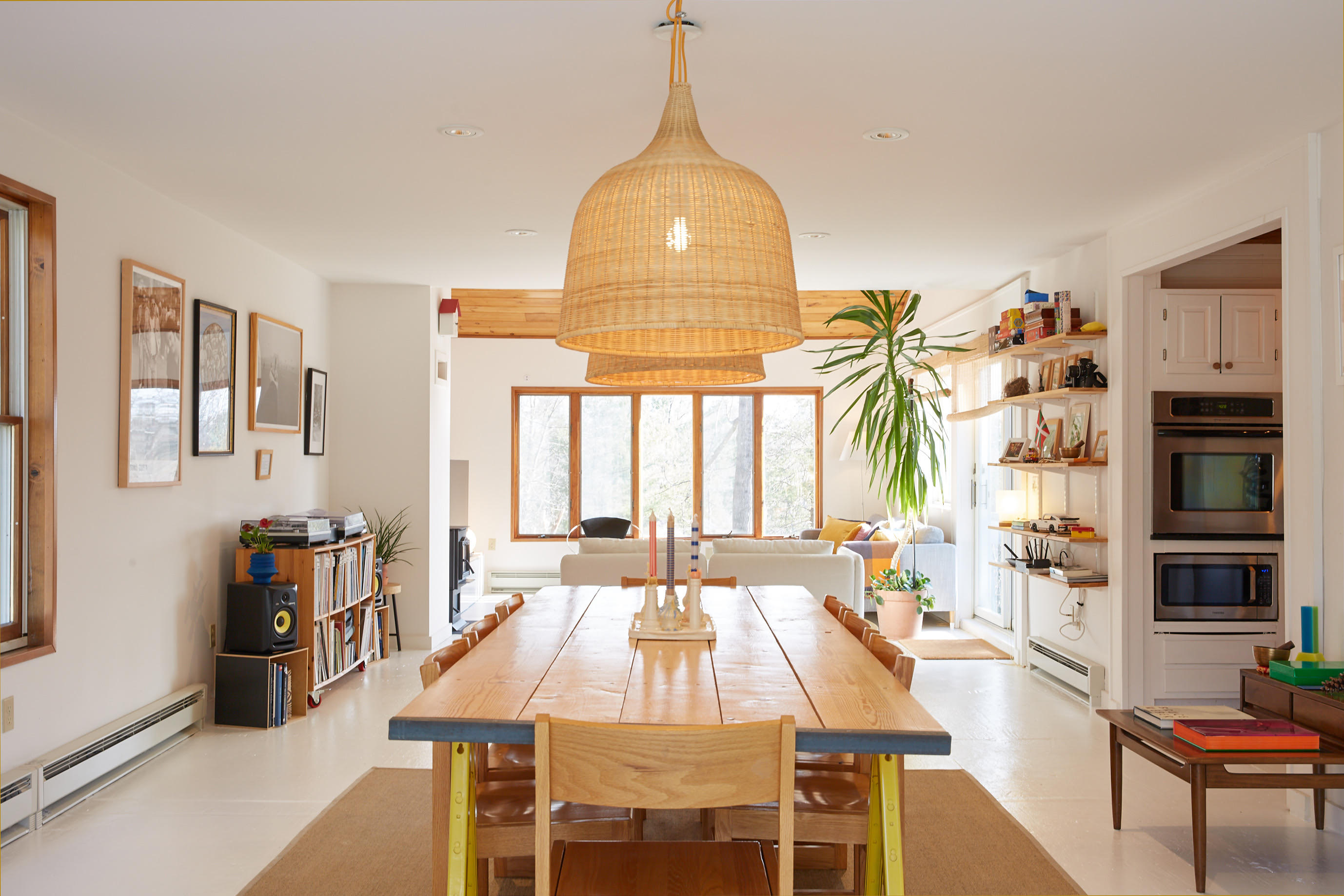 Open-concept dining area featuring a long wooden table under two large woven pendant lights, with a view into the living room and kitchen beyond. White walls and light flooring create a bright, airy feel.