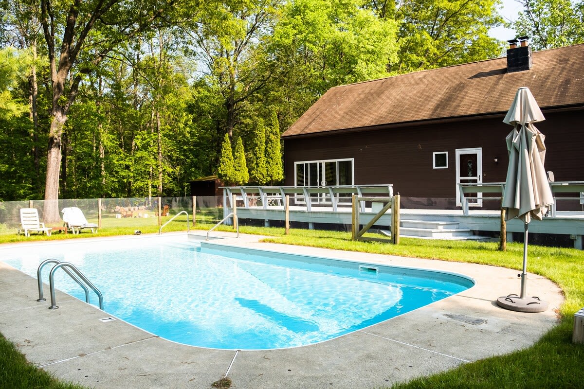 An inviting outdoor swimming pool with a concrete surround, set in a grassy yard with lounge chairs and a large umbrella. The brown-sided house with a spacious deck and mature trees frames the background.
