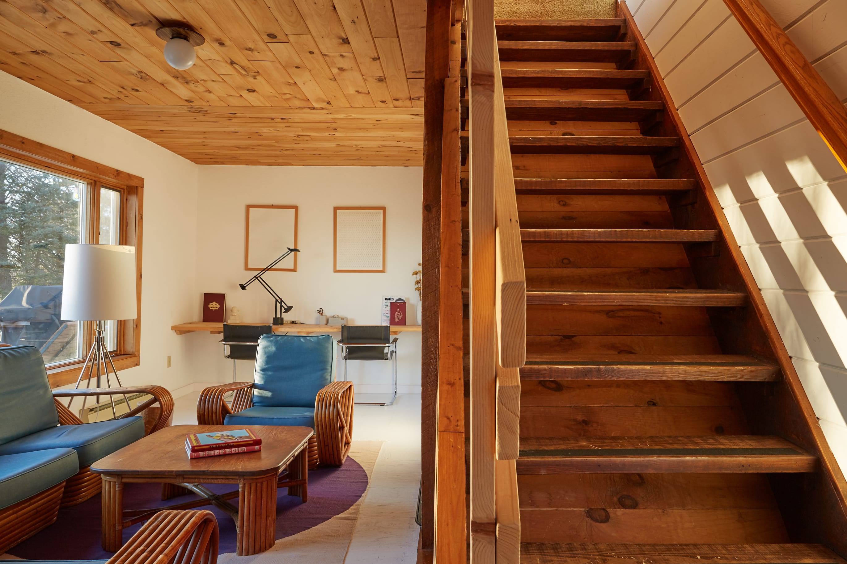 Living room with a wooden plank ceiling and large windows, featuring a wooden staircase leading upstairs. Rattan seating with blue cushions and a wooden coffee table are visible in the foreground, with a desk area in the background.