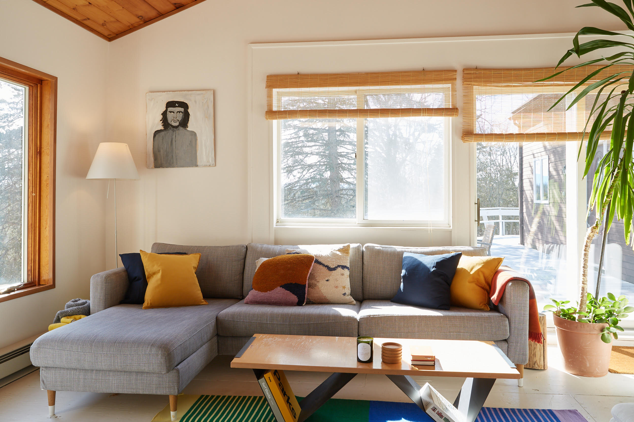 Cozy living room with a gray sectional sofa adorned with colorful throw pillows and a blanket, facing a wooden coffee table. Bamboo blinds cover windows that look out onto the deck and trees, with a floor lamp providing soft light.