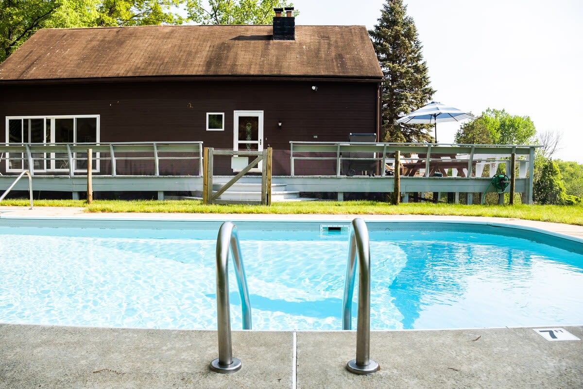 A clear blue swimming pool with prominent metal handrails in the foreground, offering a refreshing view. The brown-sided house with its deck and surrounding green landscape is visible under a bright sky in the background.