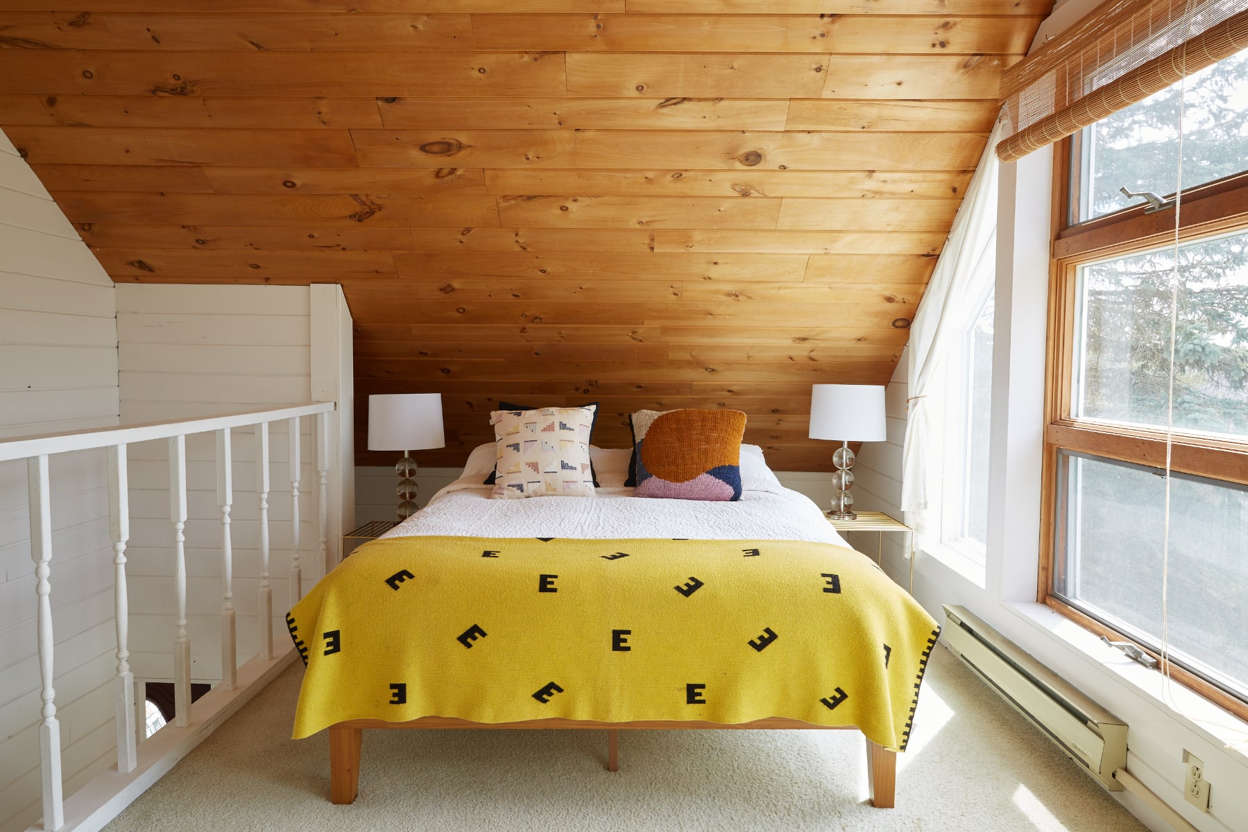 Cozy attic bedroom featuring a sloped wooden plank ceiling, white shiplap walls, and a bed with a bright yellow blanket and patterned pillows. Large windows overlook a wooded landscape, with nightstands and lamps flanking the bed.