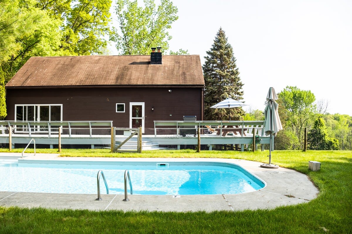 The brown-sided house with a dark roof is seen from across a sparkling blue swimming pool, surrounded by green grass and lush trees. A wooden deck with a white railing extends from the back of the house.