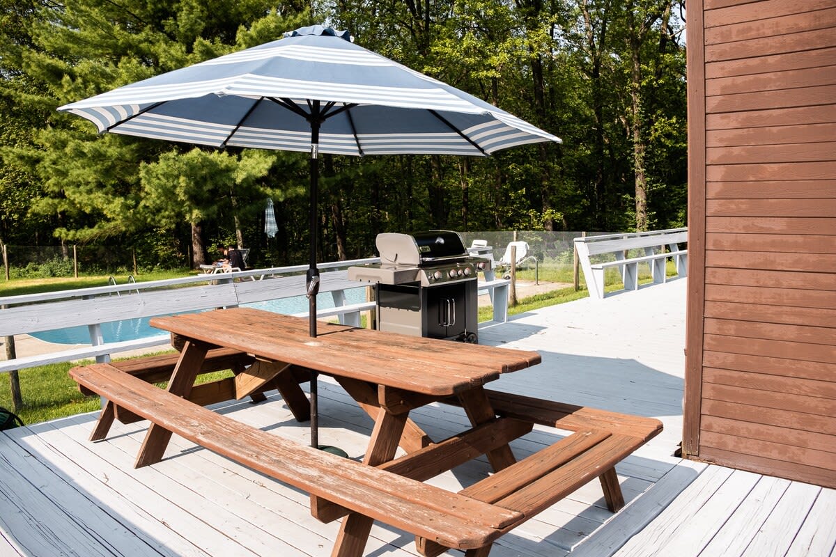 A wooden deck equipped with a picnic table and a blue-and-white striped umbrella, positioned next to a BBQ grill, offering views of the swimming pool and surrounding trees.