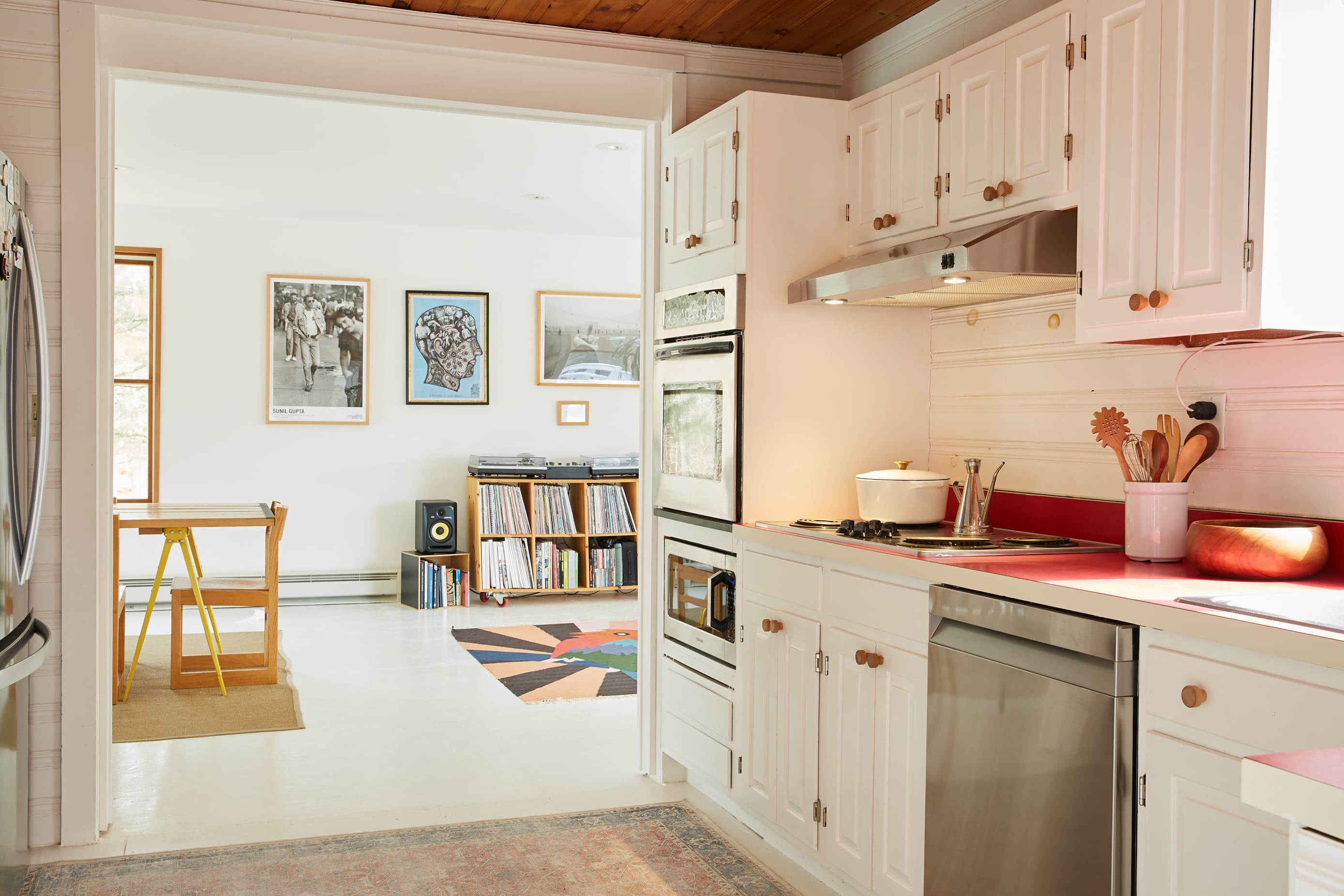 View from the kitchen into an adjacent room, revealing an open layout with white walls and light flooring. The kitchen features white cabinets, red countertops, and stainless steel appliances, with a wooden ceiling.