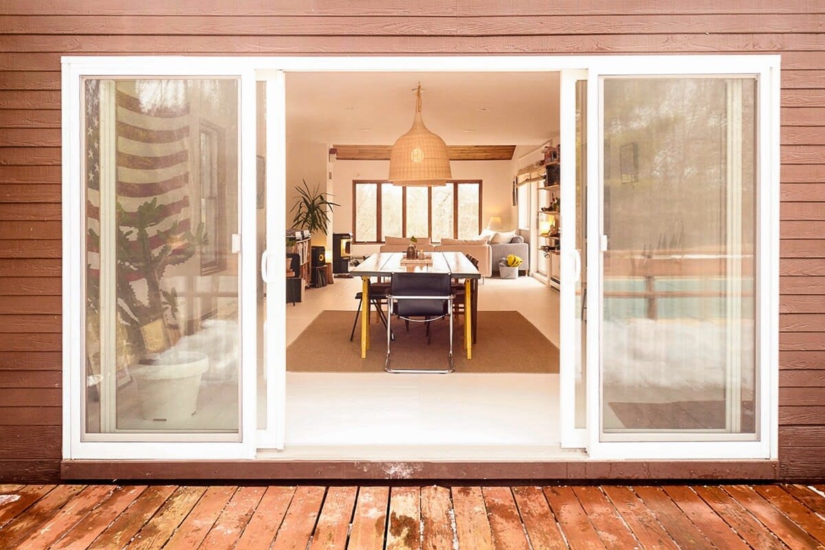 Wide sliding glass doors open from a wooden deck to reveal a bright interior dining and living area. The space features a large woven pendant light, a dining table, and a sofa, with an American flag visible through the reflection.