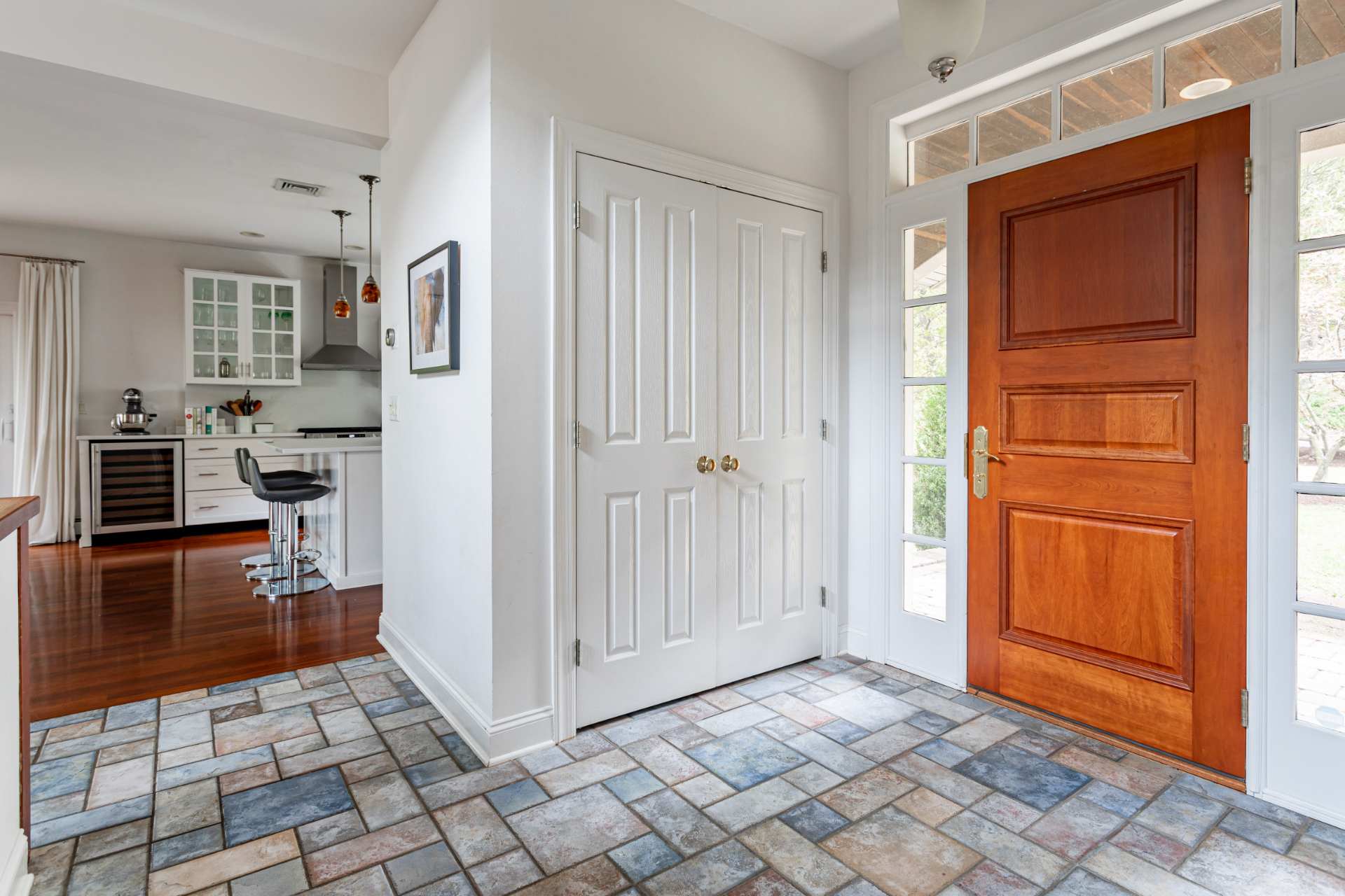 Bright foyer with multi-colored square tile flooring and a rich wooden front door, offering a glimpse into a modern kitchen with white cabinetry and a breakfast bar.