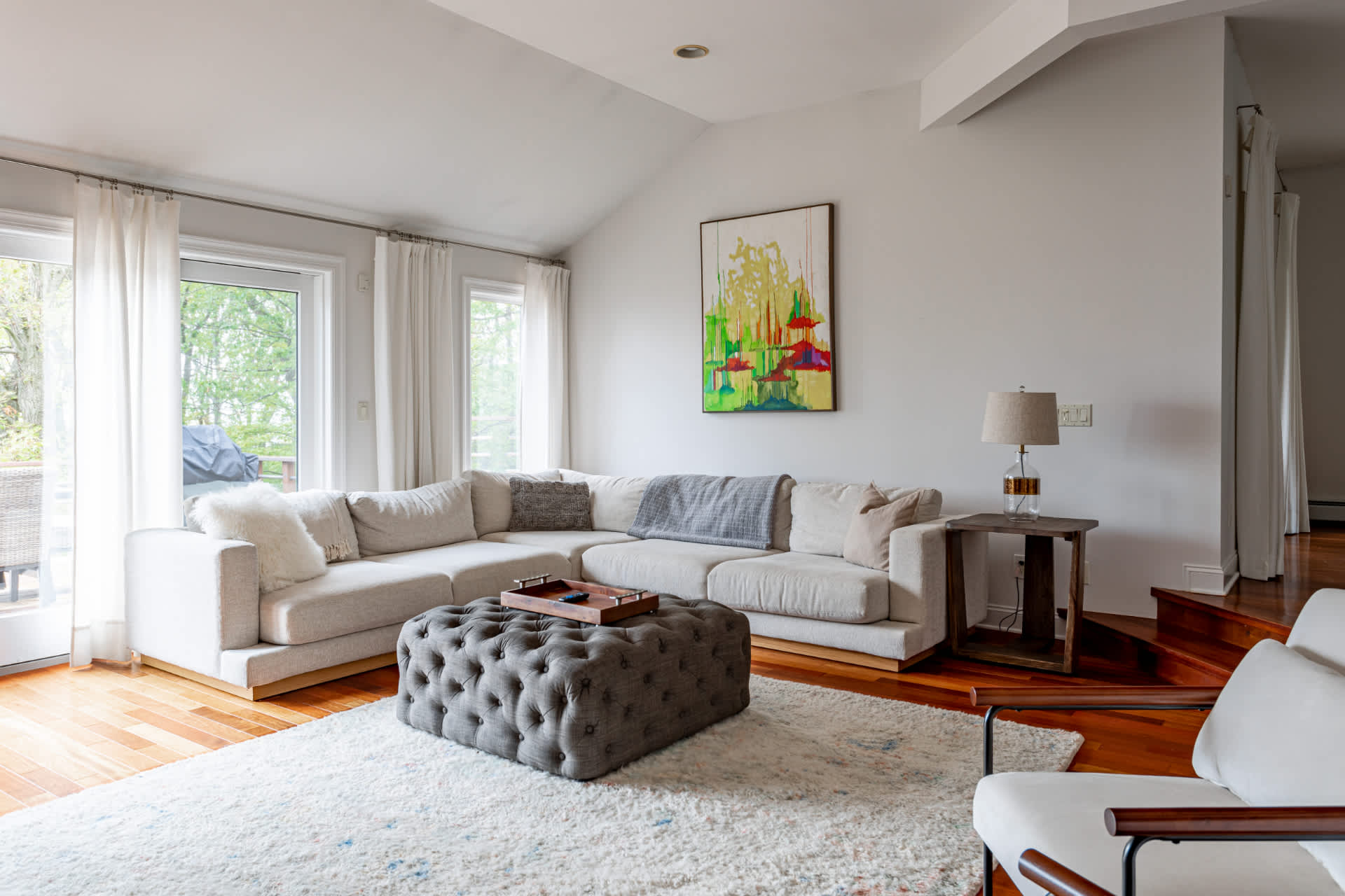 Inviting living room featuring a large light-colored sectional sofa and a gray tufted ottoman, complemented by a colorful abstract painting and a side table with a lamp.