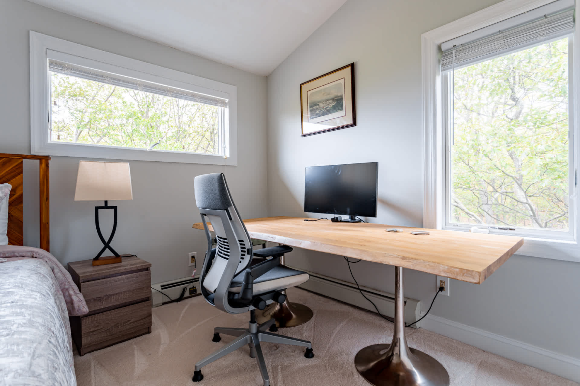 A dedicated workspace within a bedroom, featuring a light wood desk, a modern ergonomic office chair, and a computer monitor, positioned between two large windows offering views of lush trees. A bedside table and lamp are visible.