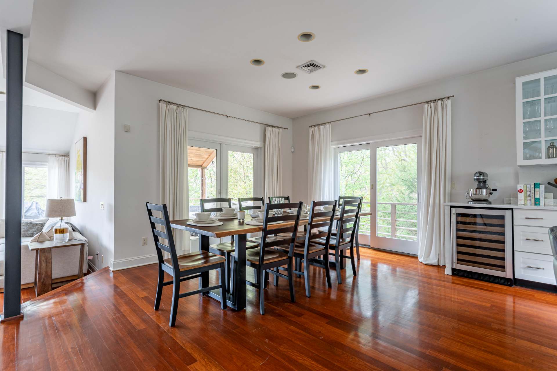 Spacious dining area featuring a long wooden dining table with black chairs, set on warm hardwood floors. Multiple glass doors with white curtains open to an outdoor deck, providing natural light and views.