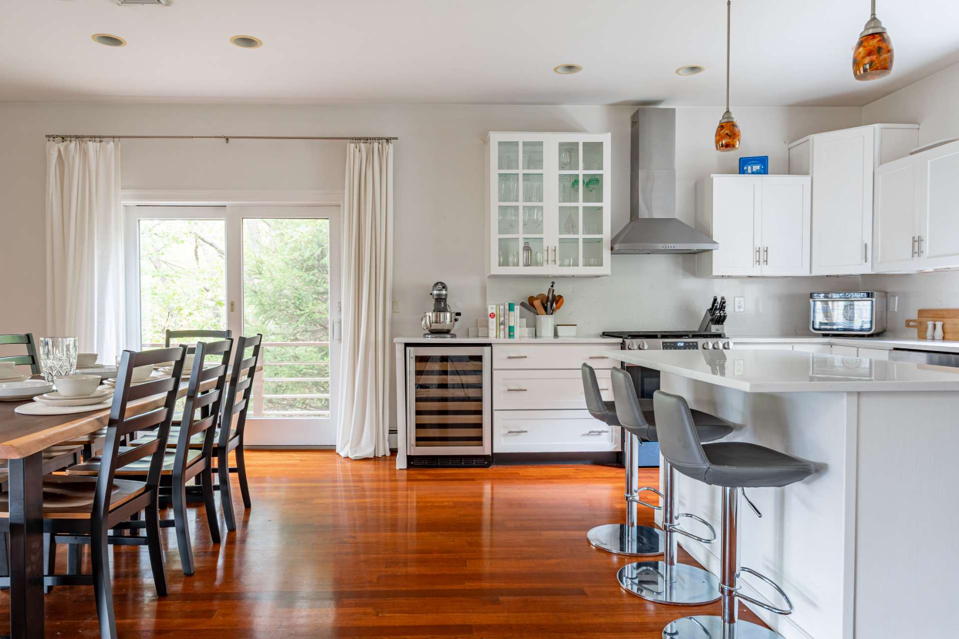 Modern kitchen with white shaker cabinets, stainless steel appliances, and a large island with bar stools, flowing into a dining area with a dark wooden table and chairs.