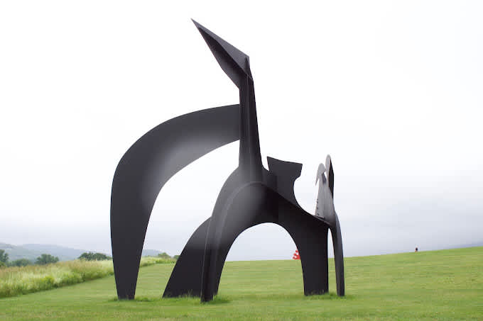 A monumental, abstract black metal sculpture with sharp angles and curved forms, standing prominently on a vast green lawn under an overcast sky, with distant hills in the background.