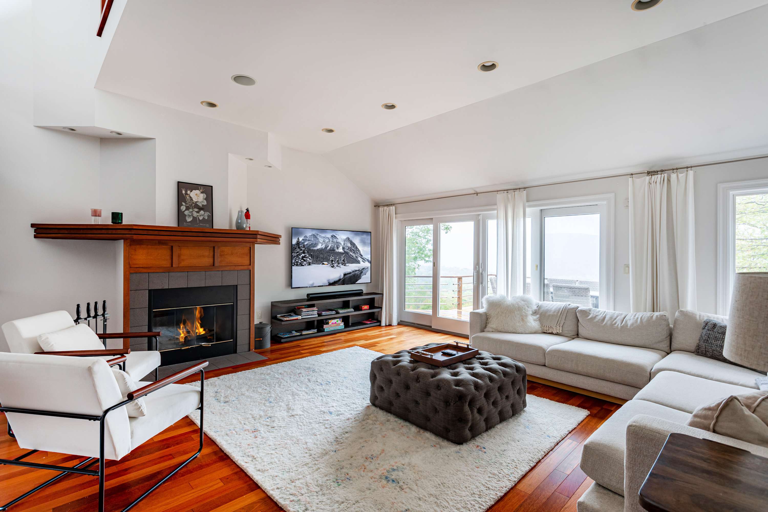 Bright living room with warm hardwood floors, a large light gray sectional sofa, a dark tufted ottoman, and a fireplace with a wooden mantel. Large glass doors with white curtains open to an outdoor space, and a flat-screen TV is mounted above a media console.