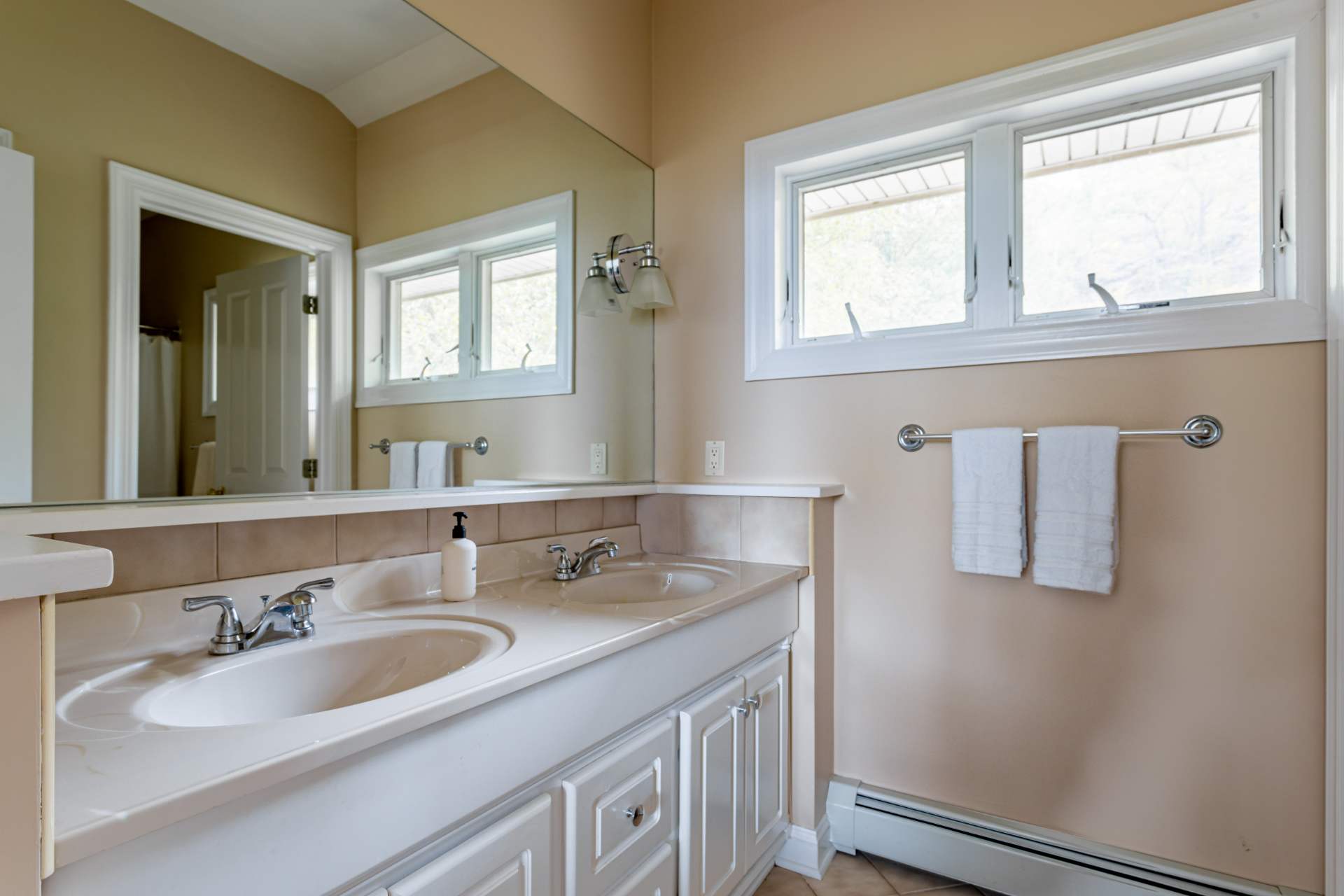 A spacious bathroom with a double vanity featuring white sinks set into a light-colored countertop, a large mirror, and two windows above a radiator. The walls are painted a warm beige, complemented by a tiled backsplash.