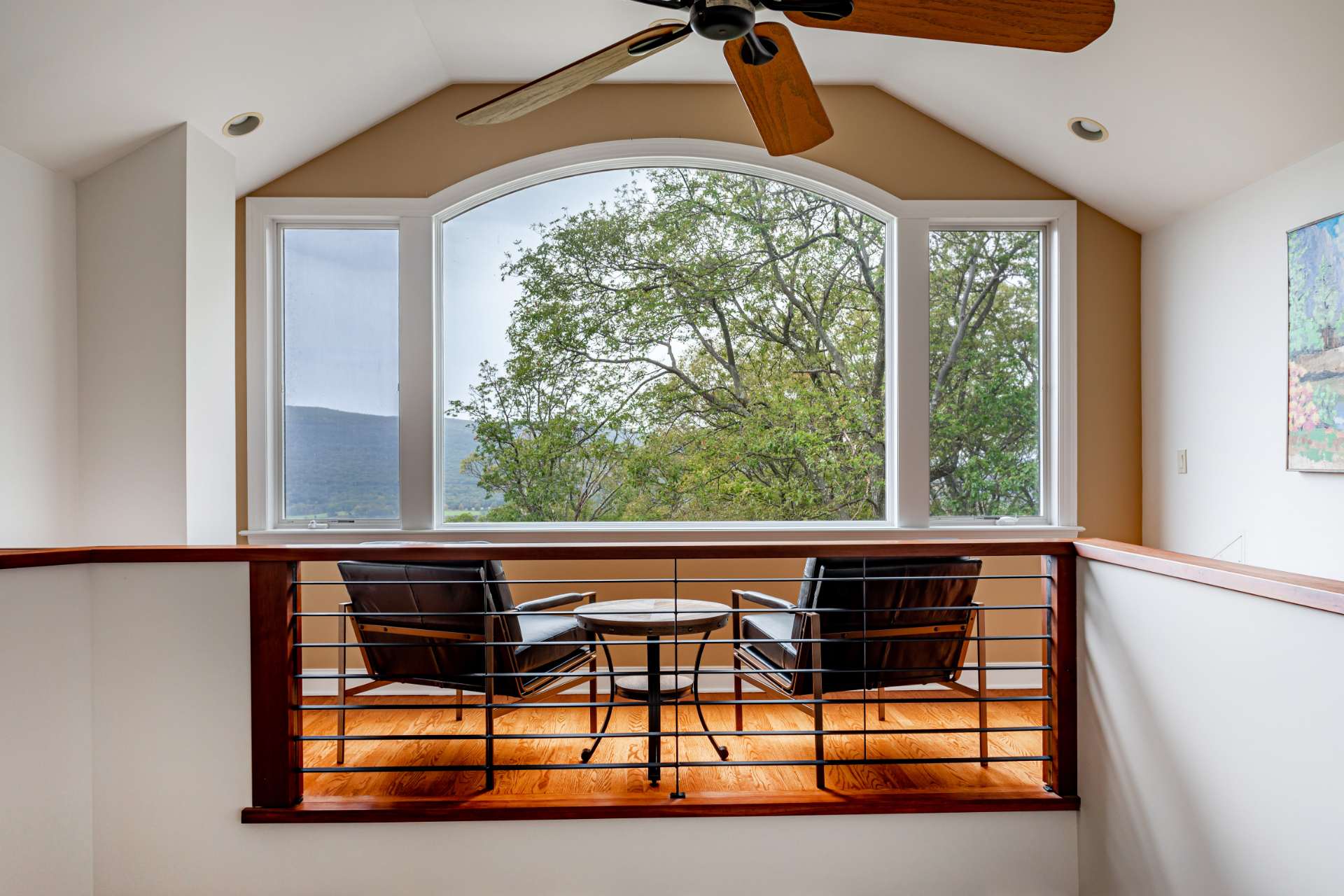 A bright loft area with a large arched window offering mountain and tree views, furnished with two black leather armchairs and a small round table. A wooden ceiling fan hangs above, and the space features hardwood floors and a modern railing.