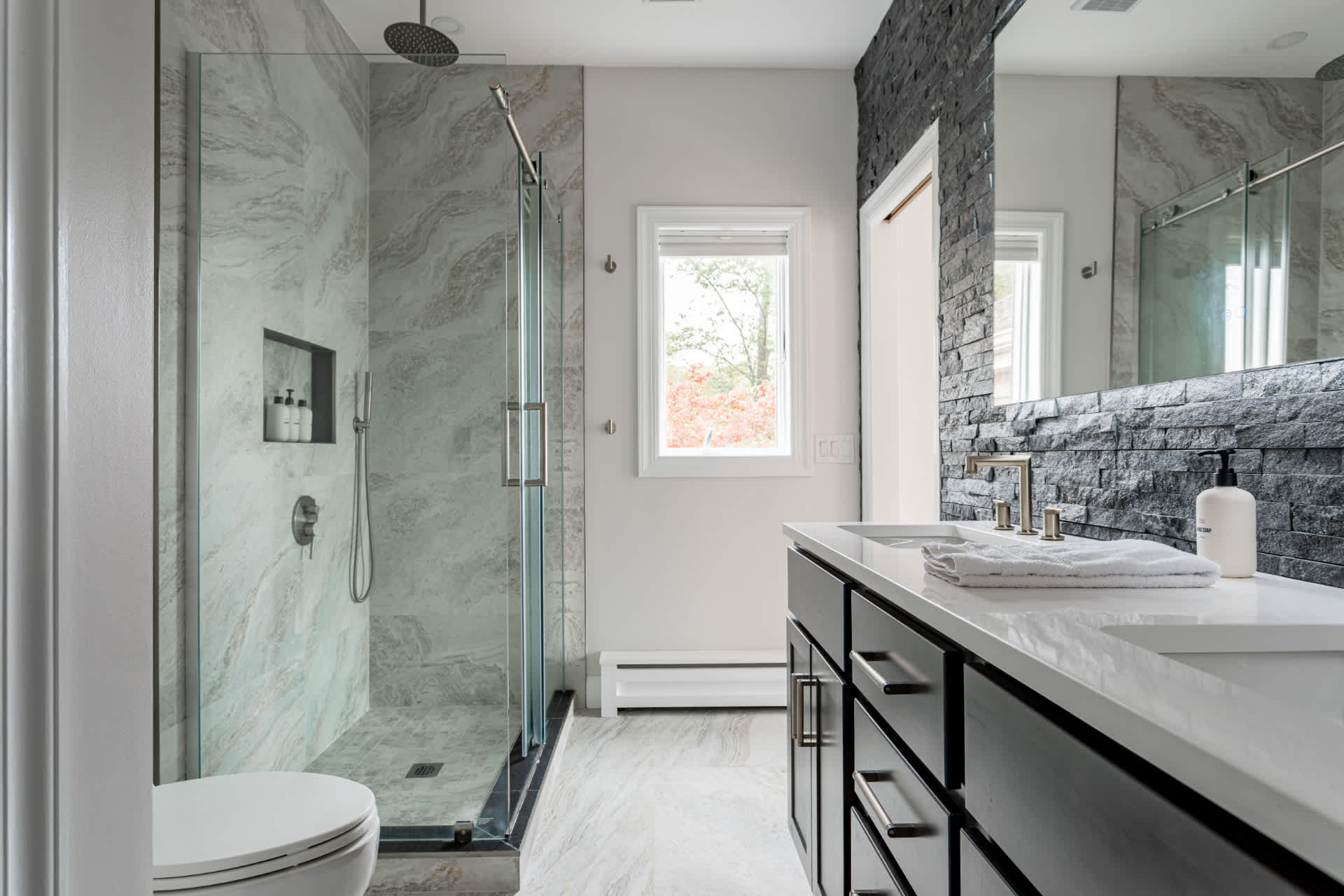 Modern bathroom with a glass-enclosed walk-in shower featuring a rain showerhead and marble-look tile, alongside a dark wood vanity with a white countertop and a window.