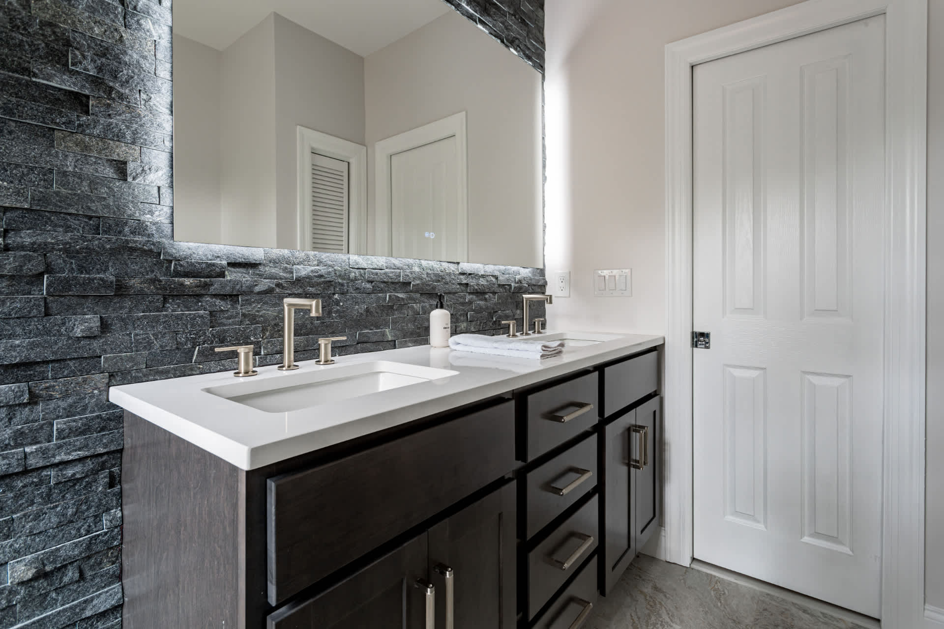 Close-up of a contemporary bathroom vanity with dual sinks, brushed nickel faucets, and a large mirror framed by a textured dark stone accent wall.