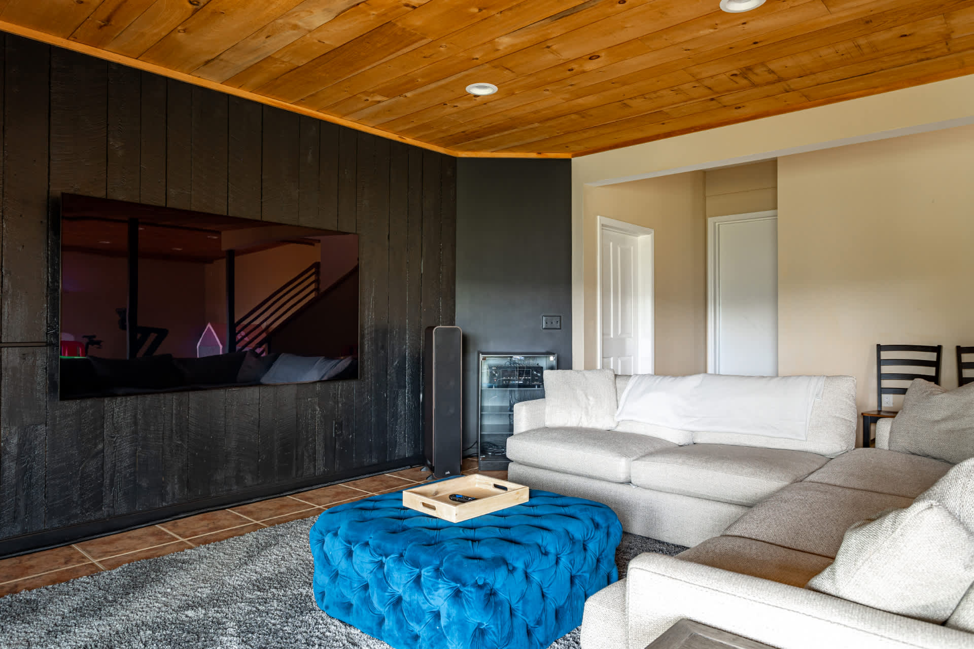Cozy media room with a large wall-mounted TV against a dark wood paneled wall, a light gray sectional sofa, and a vibrant blue tufted ottoman. The room features a warm wooden ceiling and tiled floors, creating a relaxed atmosphere.
