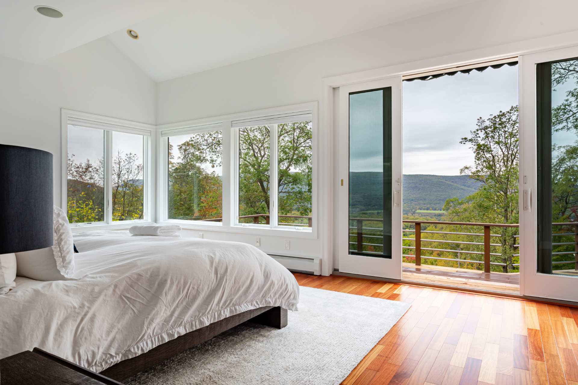 Bright bedroom featuring a white bed with a dark frame, hardwood floors, and expansive windows offering panoramic mountain and valley views. A sliding glass door opens to an outdoor deck.