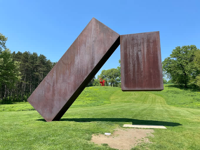 A large, abstract outdoor sculpture made of rusted metal, featuring two rectangular blocks, one tilted and the other suspended, set on a vibrant green lawn under a clear blue sky.